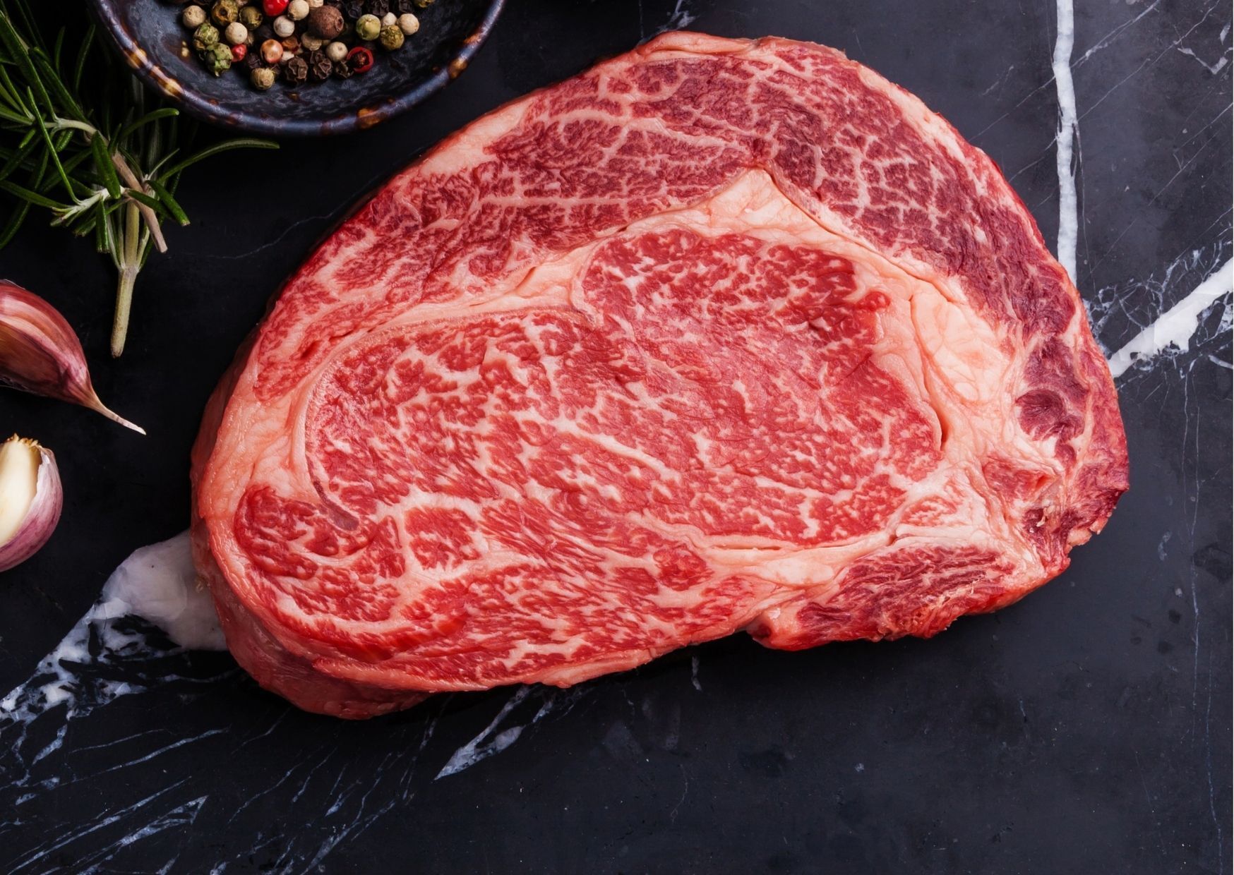 Close-up of a raw ribeye steak with heavy marbling, set on a dark surface with spices and garlic.