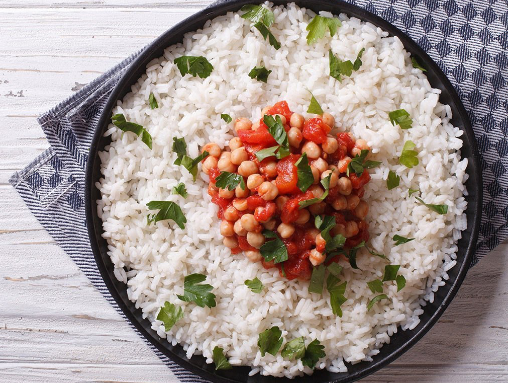 White rice with a tomato and chickpea topping, garnished with green herbs, served on a black plate.