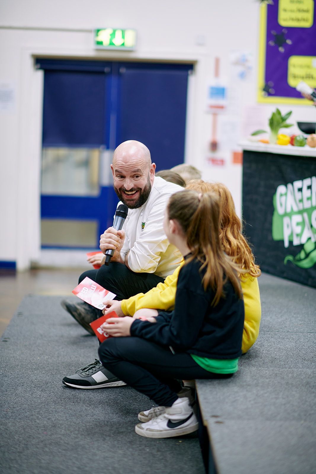 Man speaking into a microphone to two children, seated on a stage, at an event.