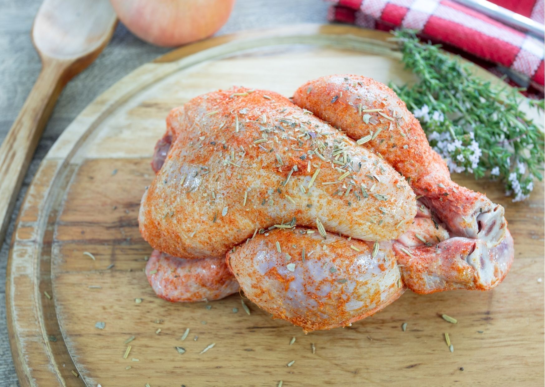 Seasoned chicken pieces on a wooden cutting board with onion, herbs, and a red checkered cloth.