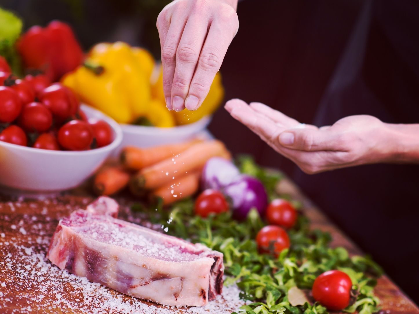 Person salting raw steak with vegetables and ingredients in the background.