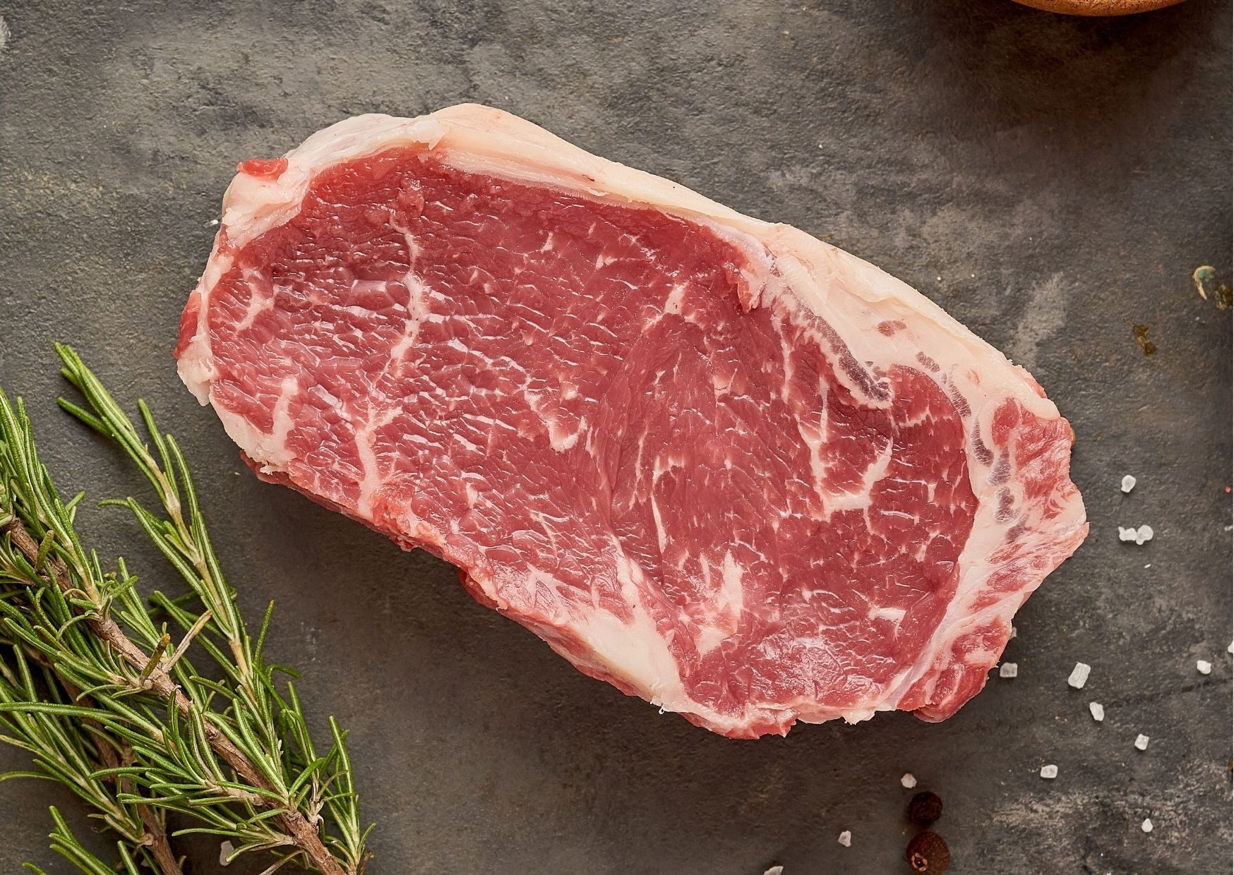 Raw New York strip steak, red with marbling, rosemary, and salt.