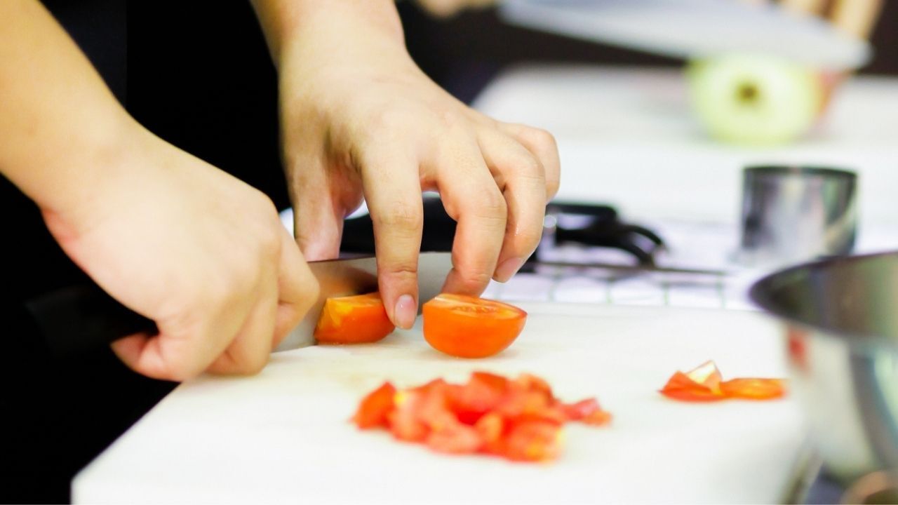 Hands slicing a tomato on a white cutting board, with diced tomatoes, and a partially seen stove and pot.