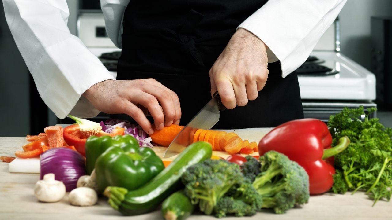 Chef cutting a carrot on a cutting board surrounded by various colorful vegetables in a kitchen.