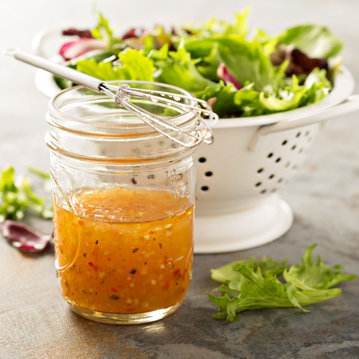 Jar of dressing with whisk next to a colander of salad greens.