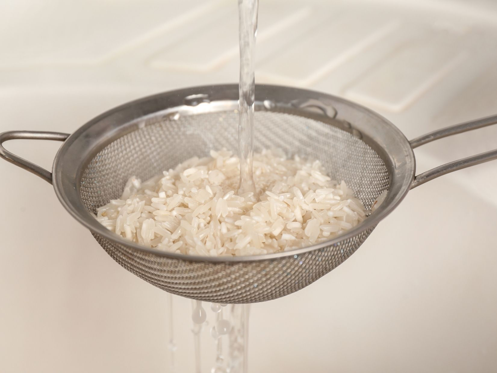 Rice being rinsed in a metal mesh strainer under running water in a sink.