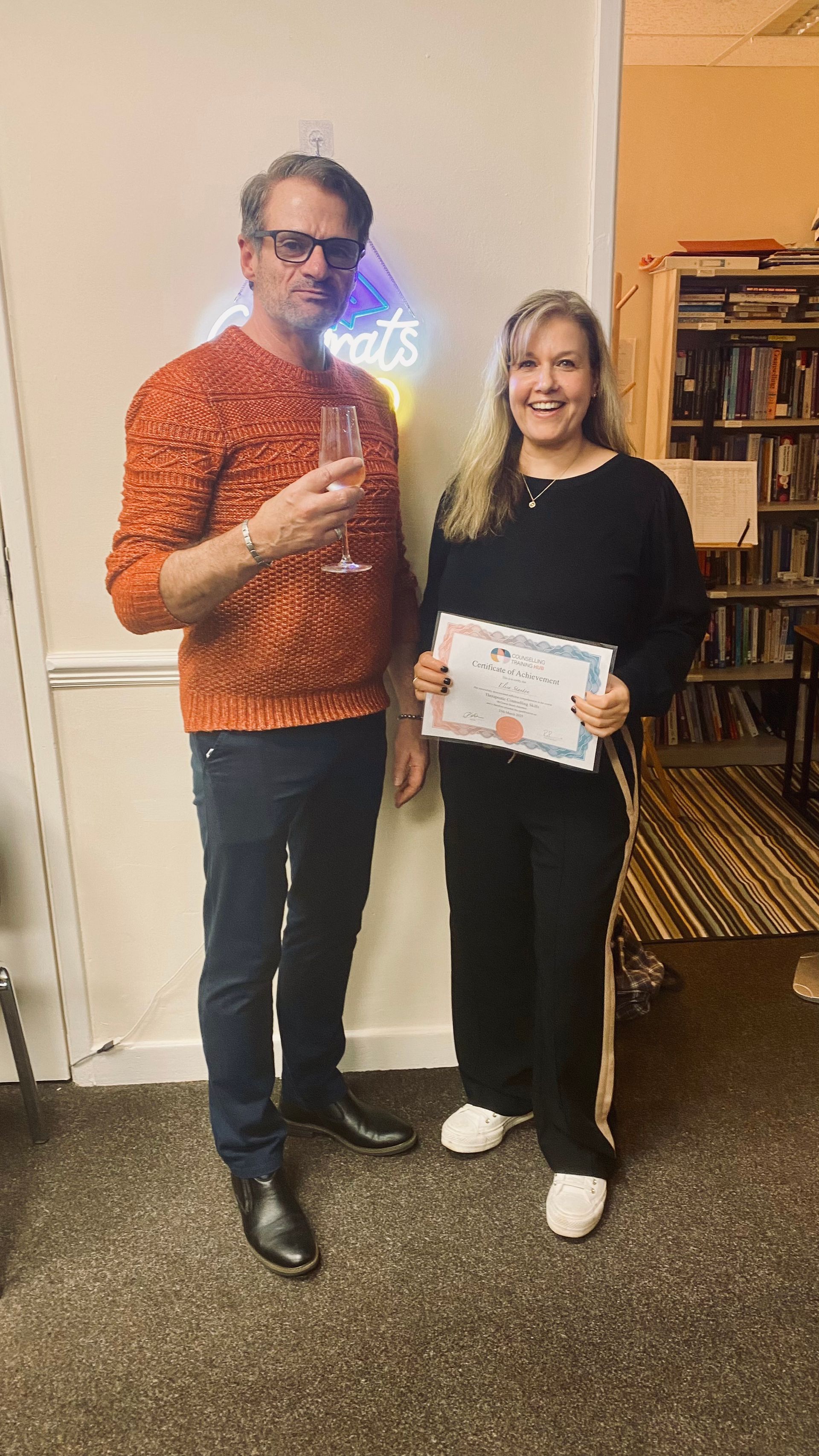 Man in orange sweater and woman in black outfit holding a certificate, celebrating; bookshelf in background.