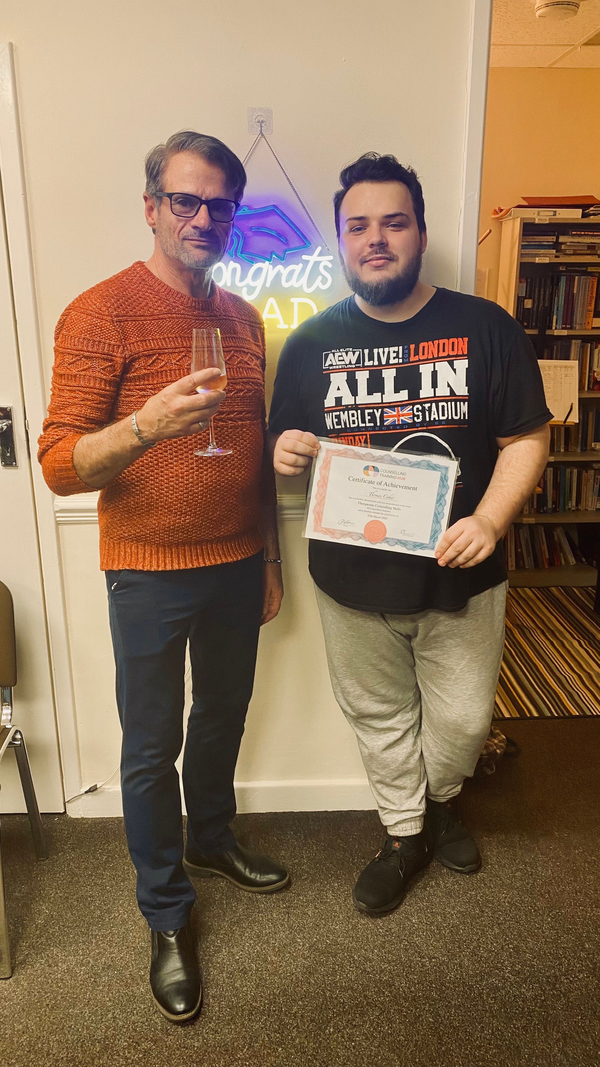 Two men pose indoors. One holds a champagne glass, the other a certificate. A neon sign says 