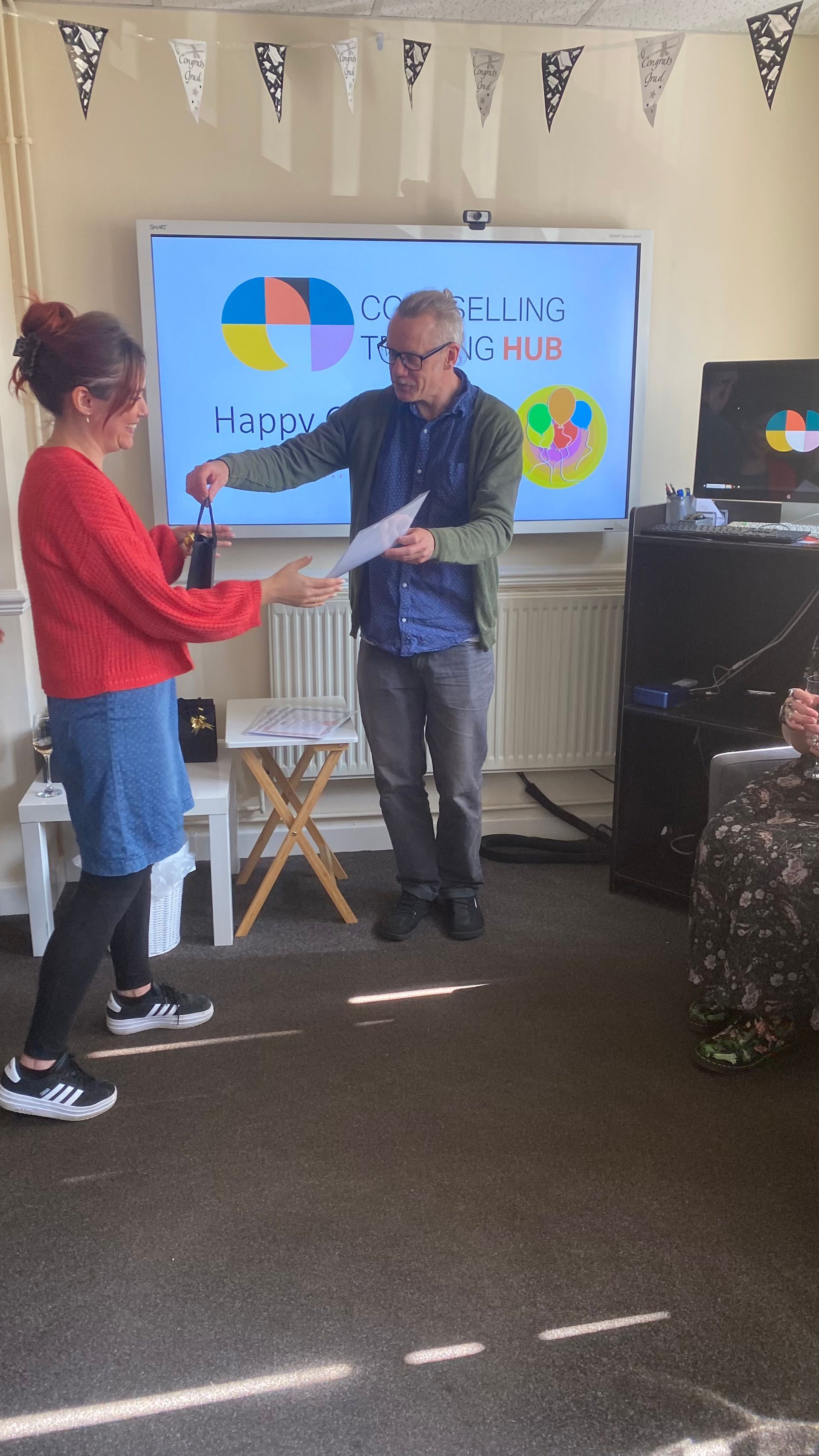 Woman receives a document from a man, in front of a presentation screen, bunting overhead.