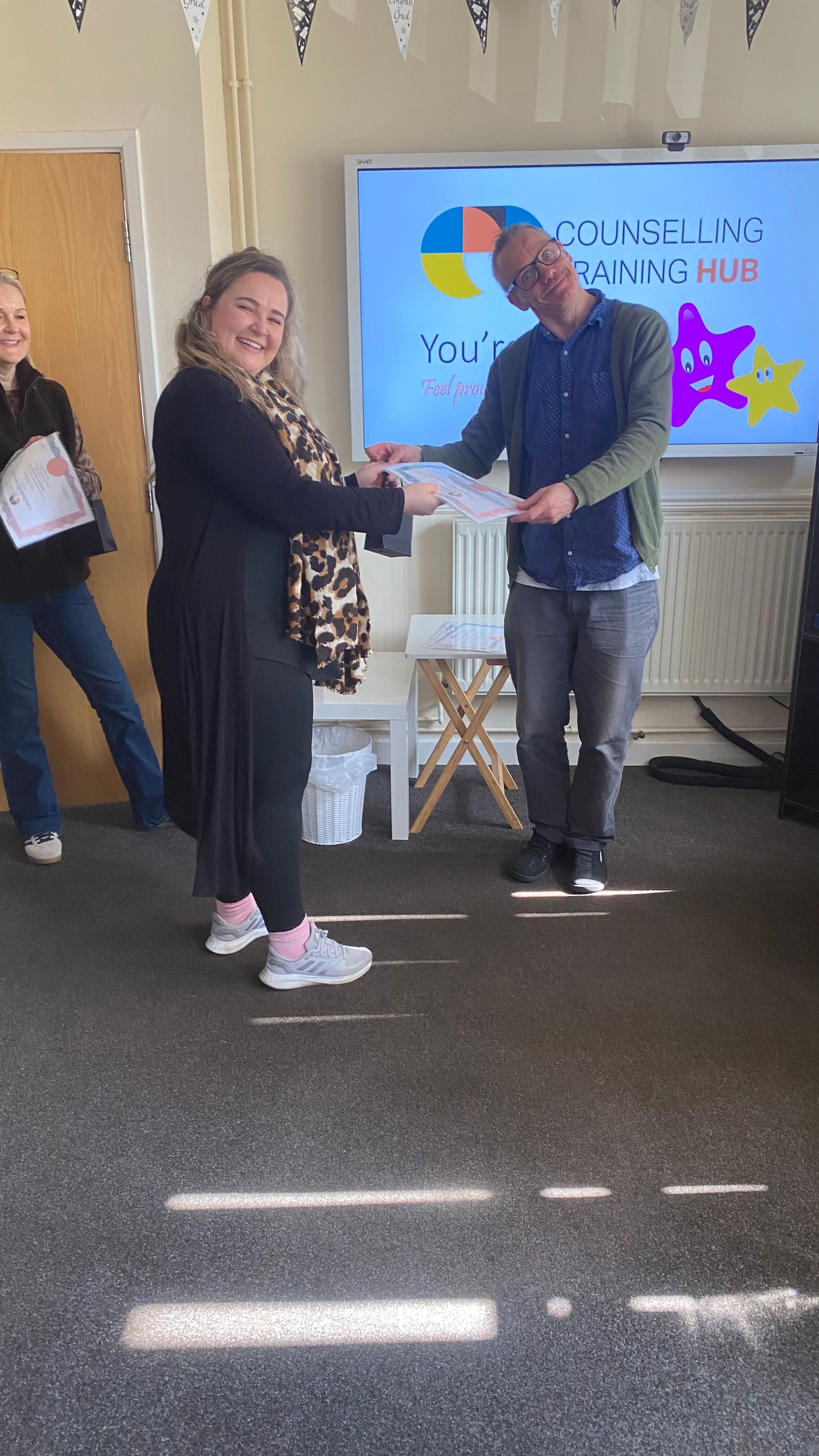 Woman receiving a document from a man, in front of a screen with a logo, indoors.