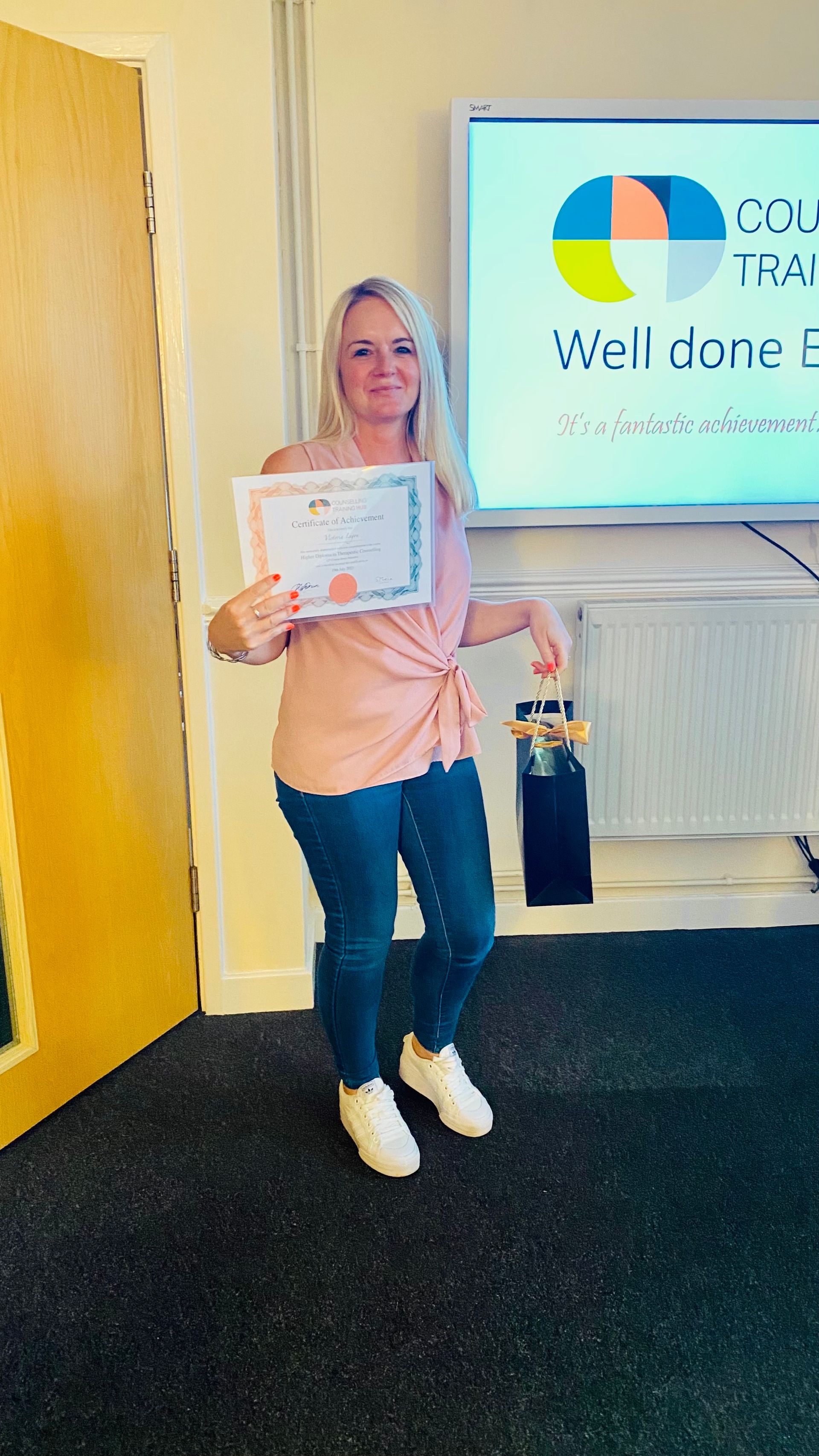 Woman in jeans and pink top holding a certificate, smiling, in front of a screen that says 
