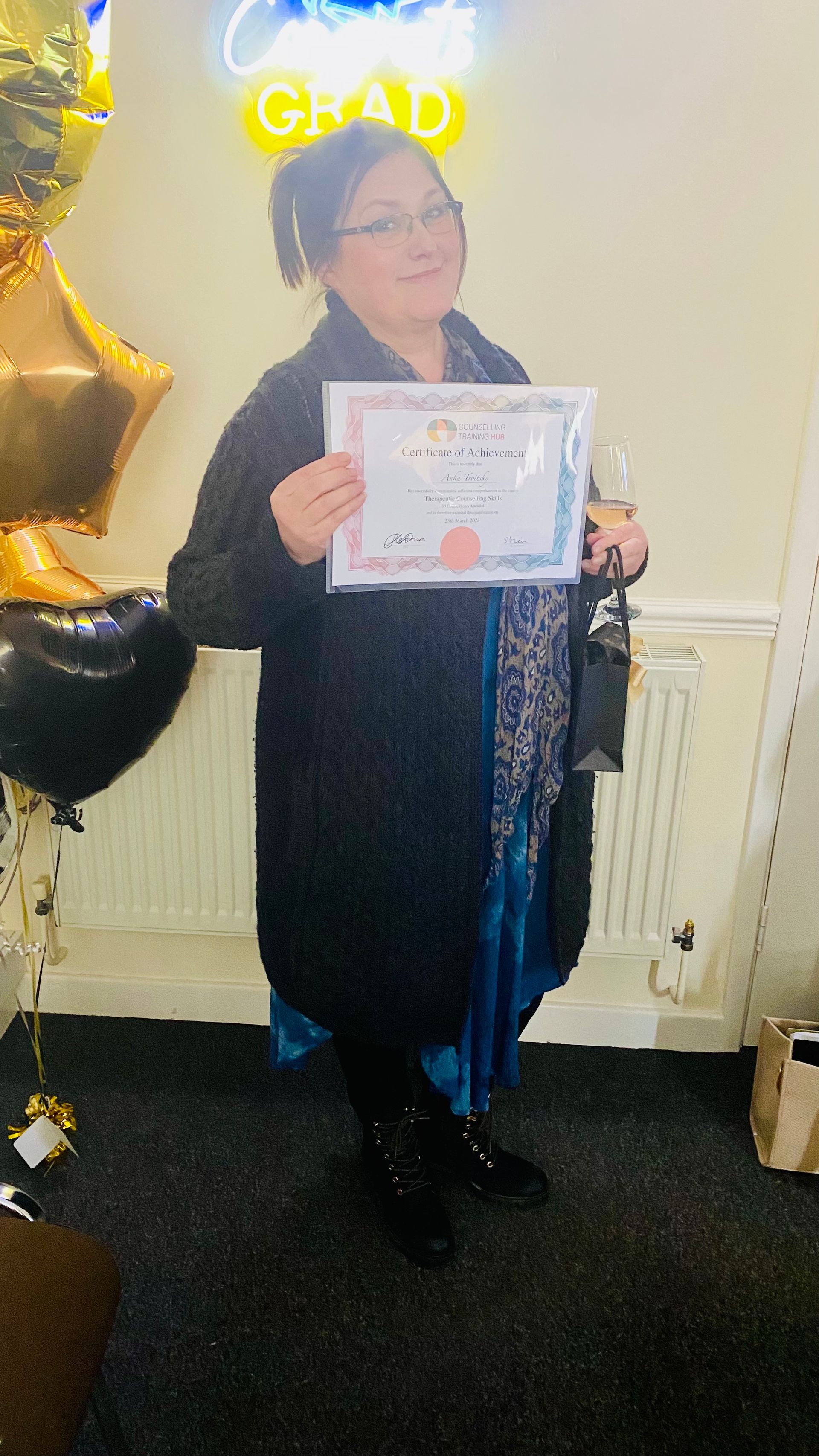 Woman in black coat, holding certificate, smiling at graduation party with balloons.