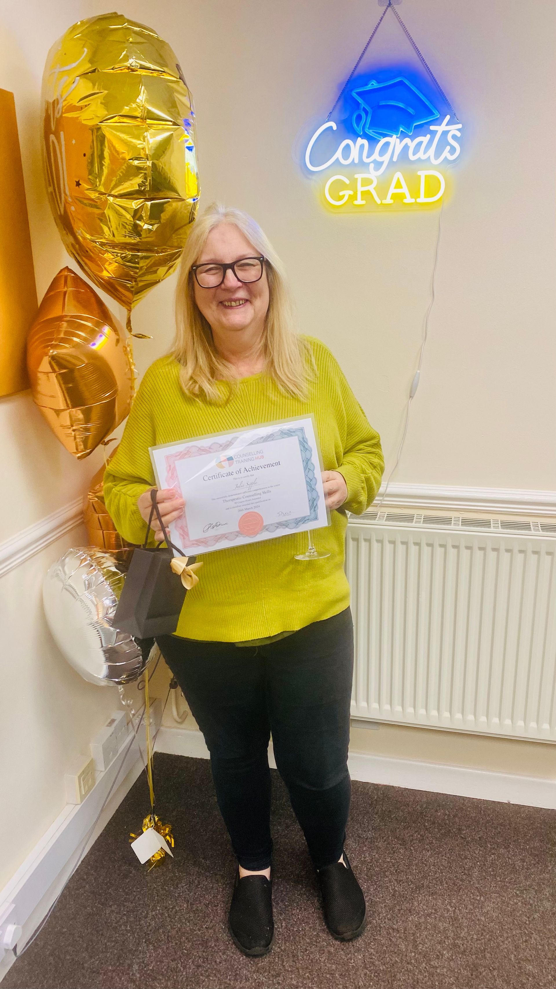 Woman in glasses holds a certificate, celebrating graduation, with balloons and a 