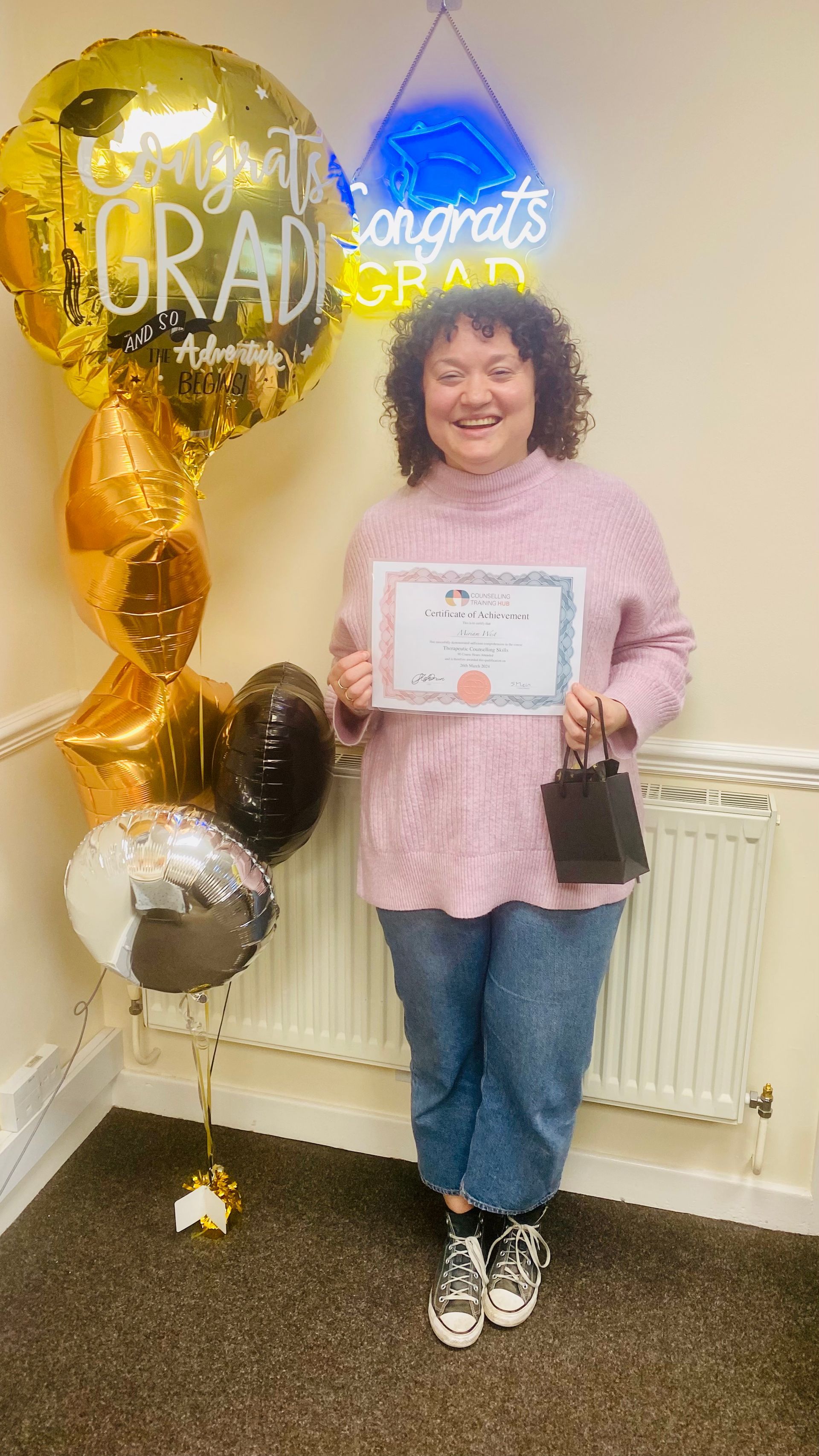 Woman holds a certificate, smiling, standing by balloons that say “Congrats GRAD.”