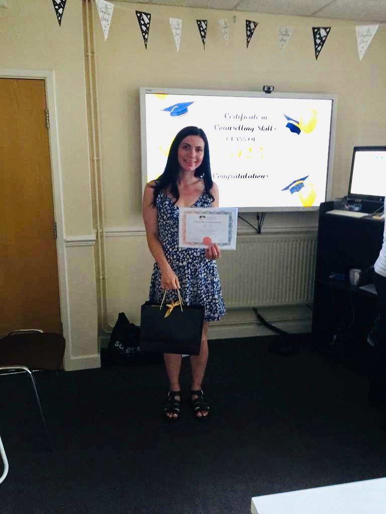 Woman in a blue dress holding a certificate and a bag, standing in front of a whiteboard.