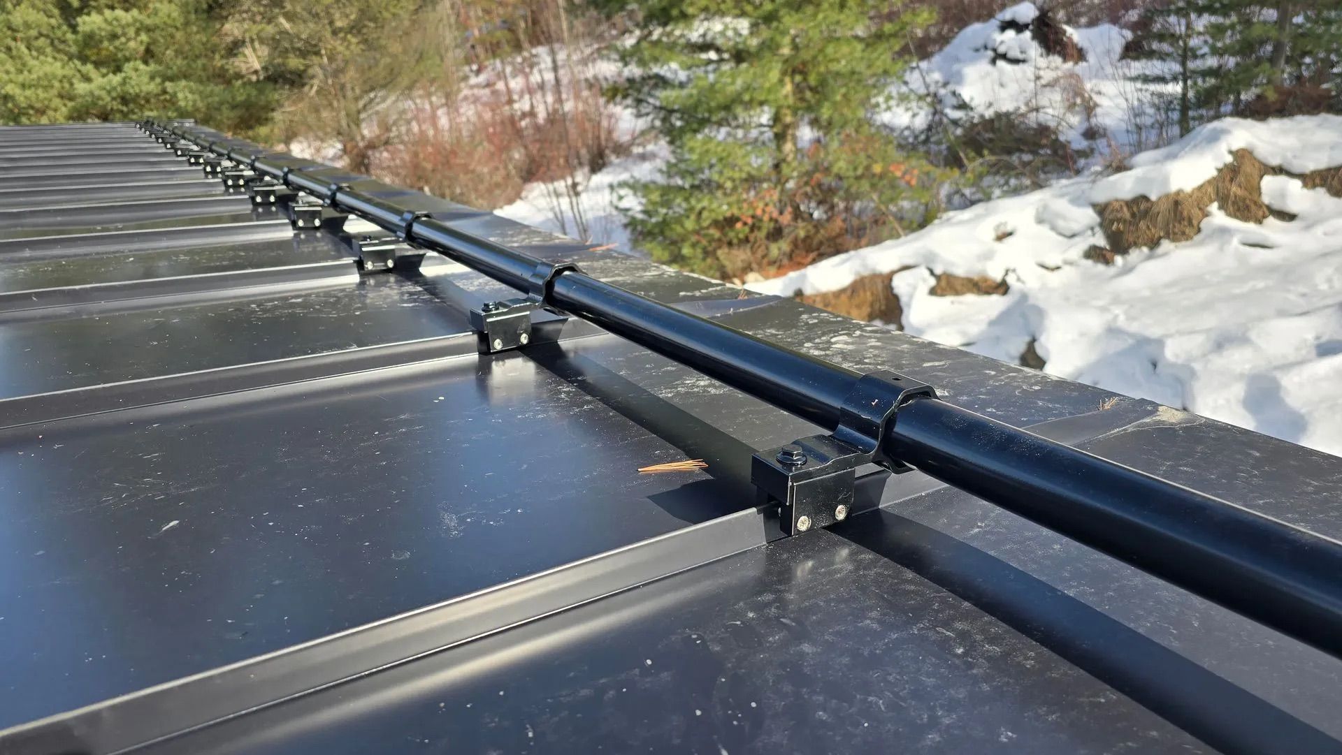 Black solar panels with mounting rails beside snow on a sunny wooded hillside