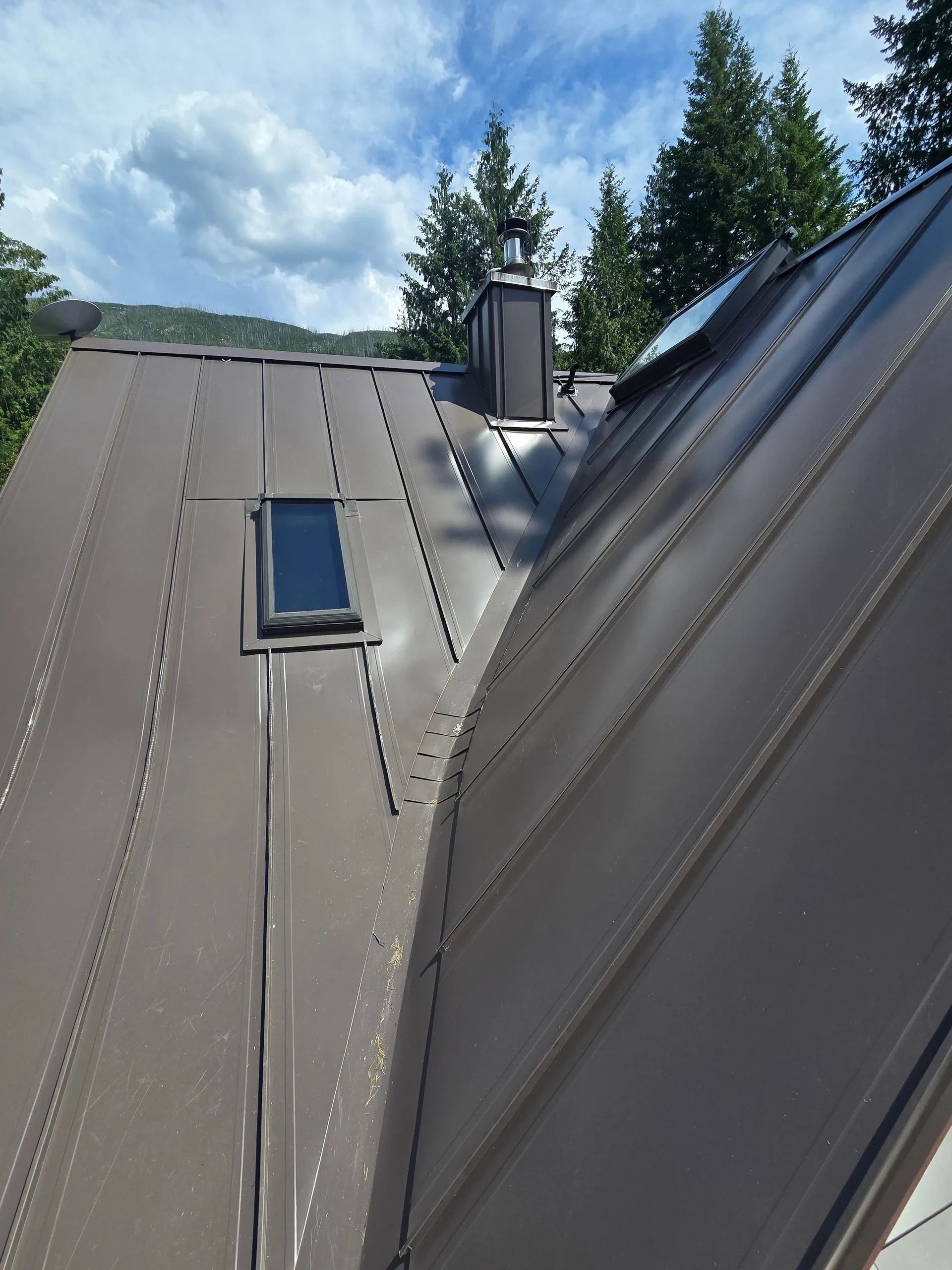 Metal roof with skylight and chimney under a cloudy sky, viewed from a sloped rooftop