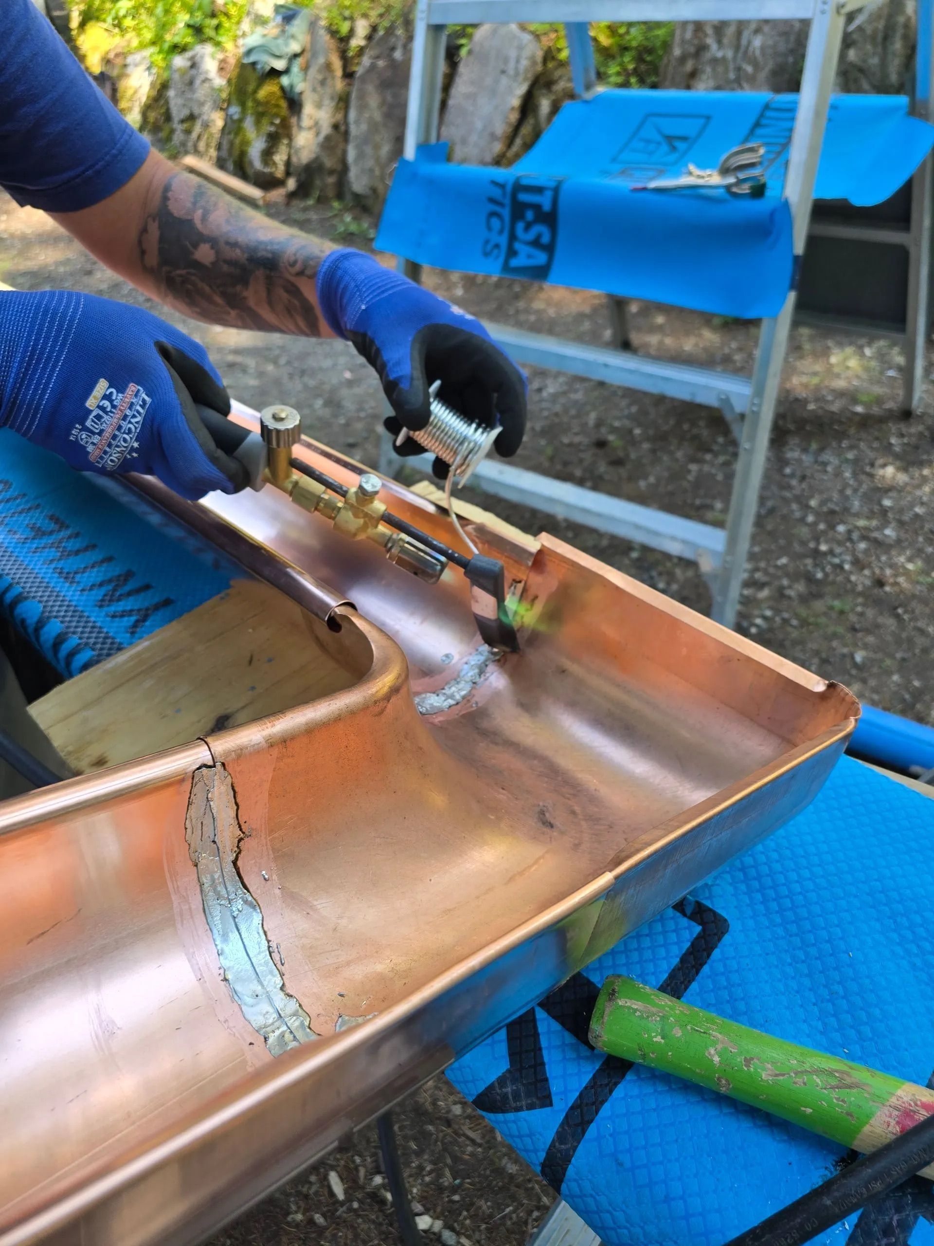 Worker welding a copper gutter outdoors beside a ladder and tools