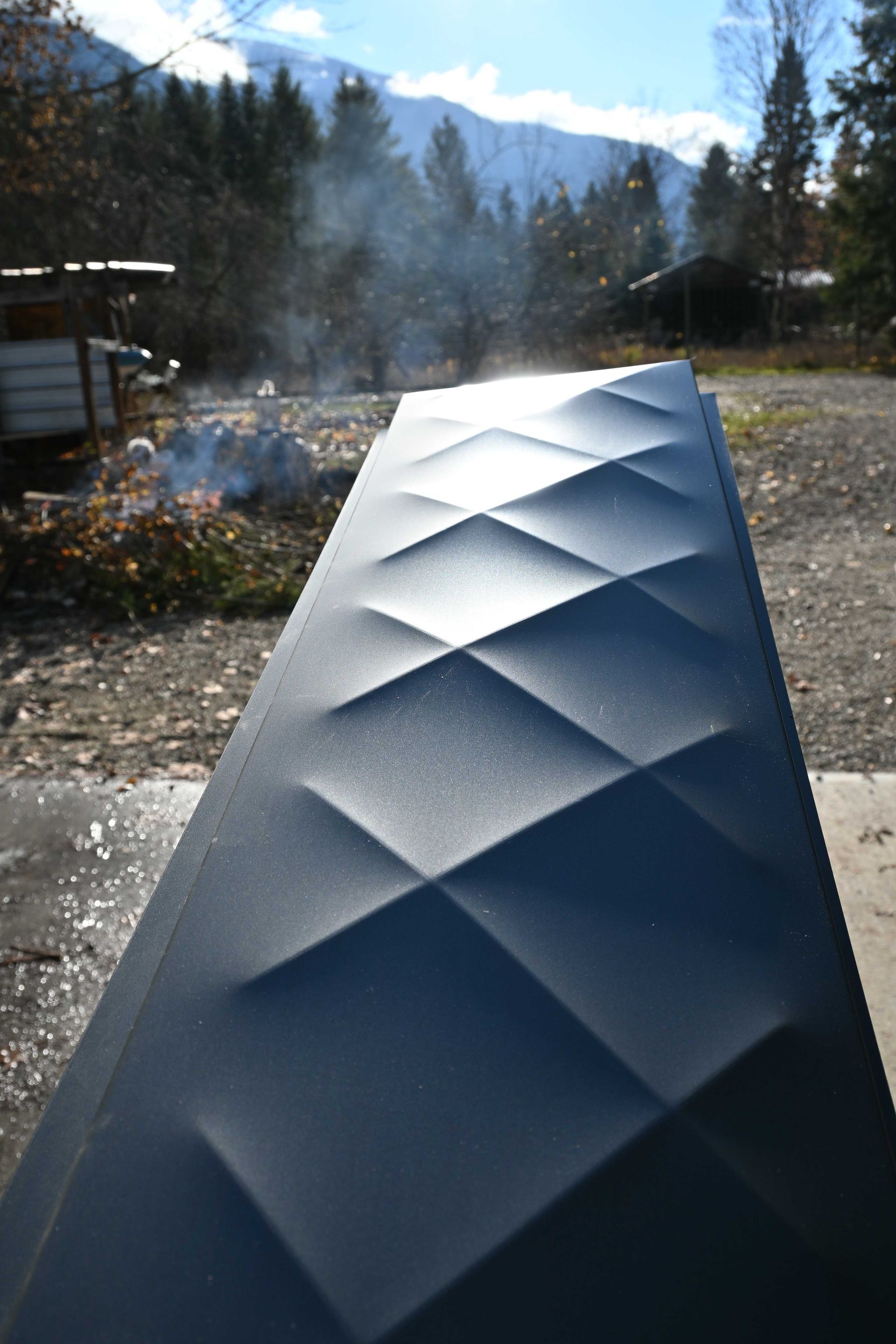 Silver diamond-textured metal panel outdoors in sunlight with mountains and trees in the background