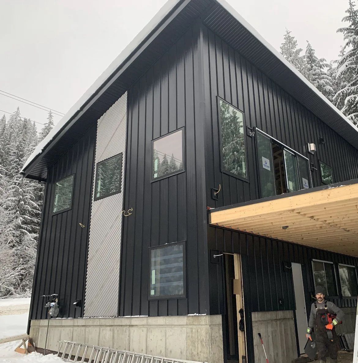 Black modern cabin with large windows and a wood overhang in a snowy setting