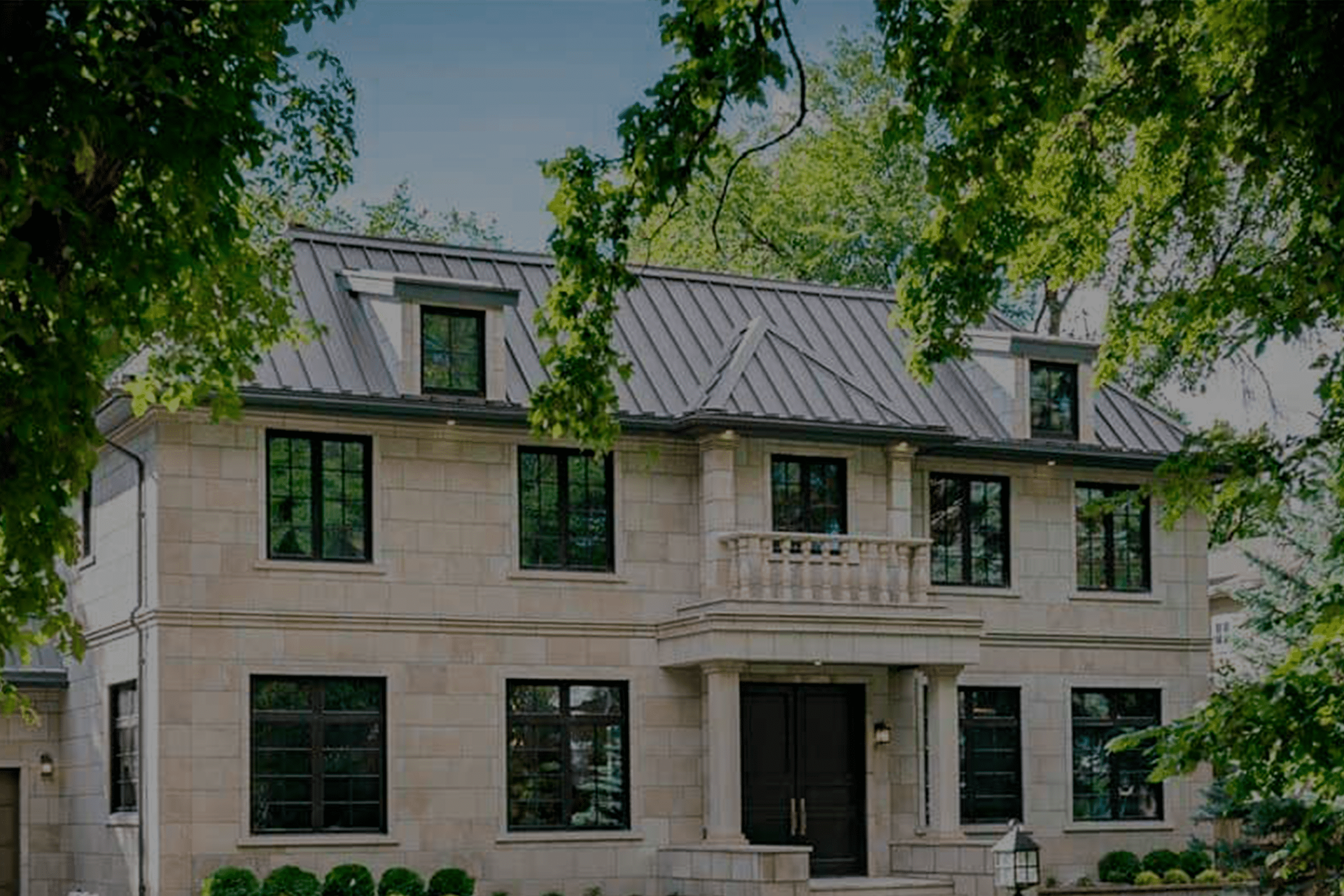 Large stone house with a gray metal roof, black-trimmed windows, and trees framing the front yard.