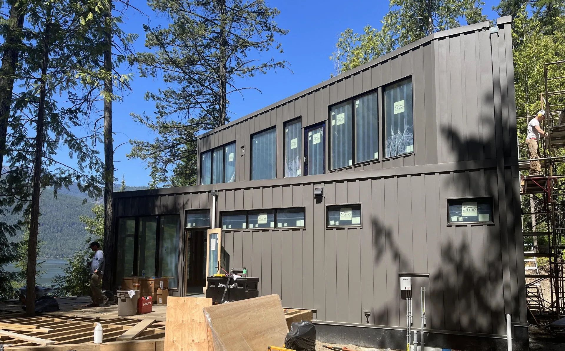 Modern gray two-story house with large windows in a wooded hillside setting, deck and patio chairs outside