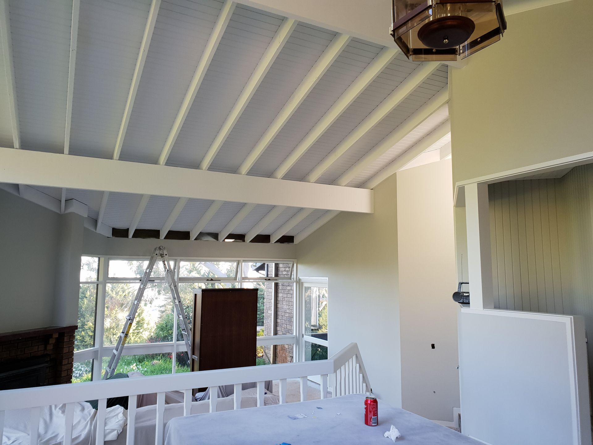 Covered porch with white ceiling beams, railing, windows, and a red bottle on a table