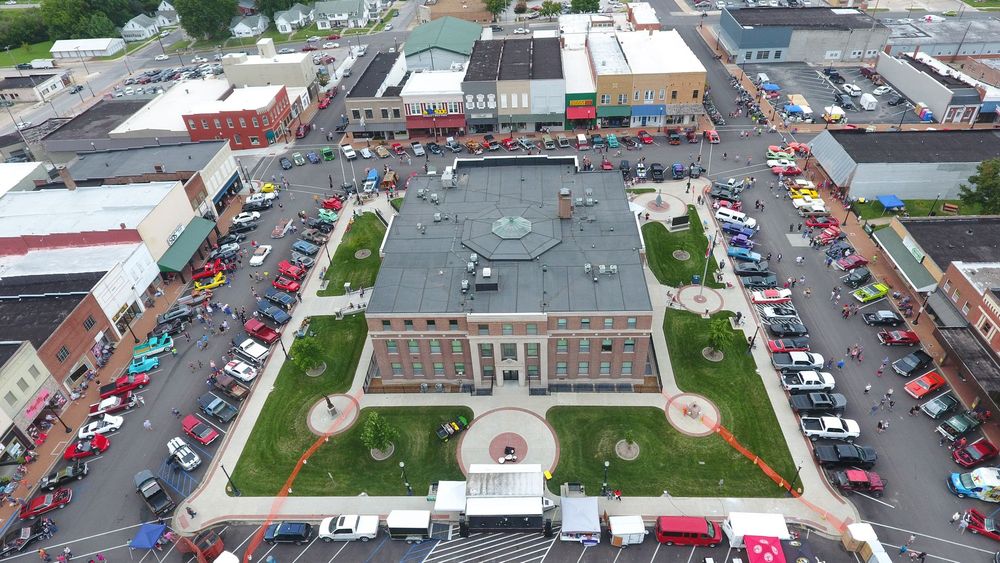 Aerial view of a town square with a brick building at its center surrounded by cars; event in progress.