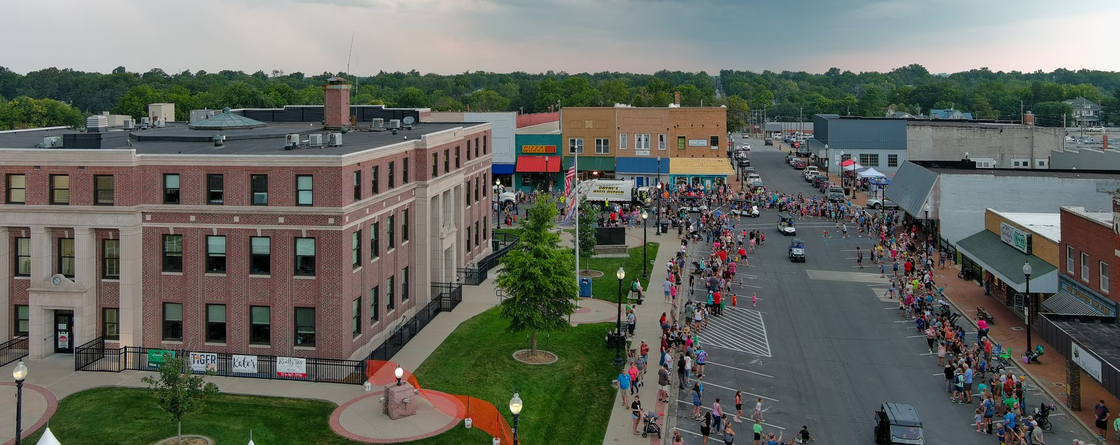 Aerial view of a busy street fair beside brick buildings and tents, with crowds lining the road.