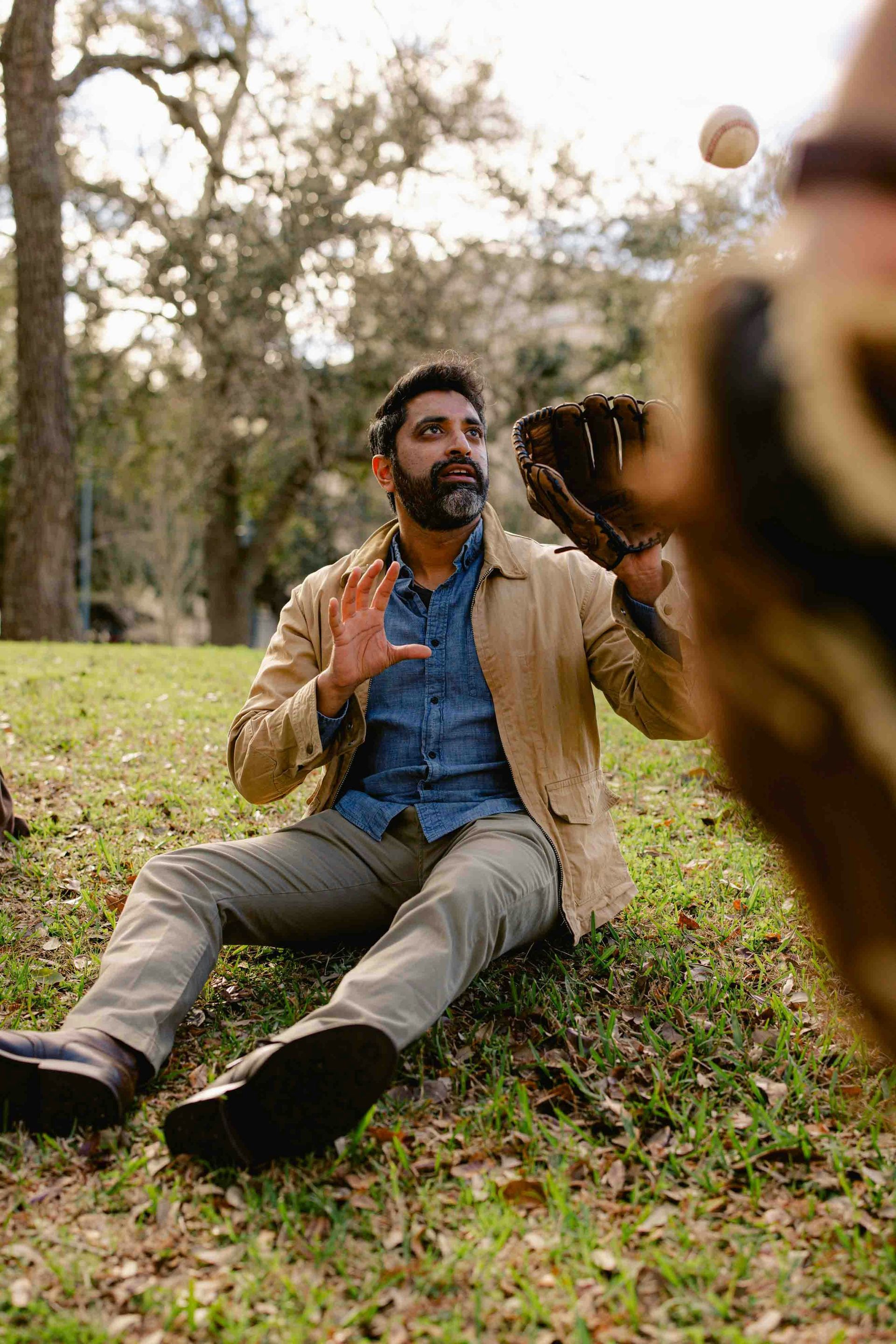 A man is sitting on the ground in a park catching a baseball.