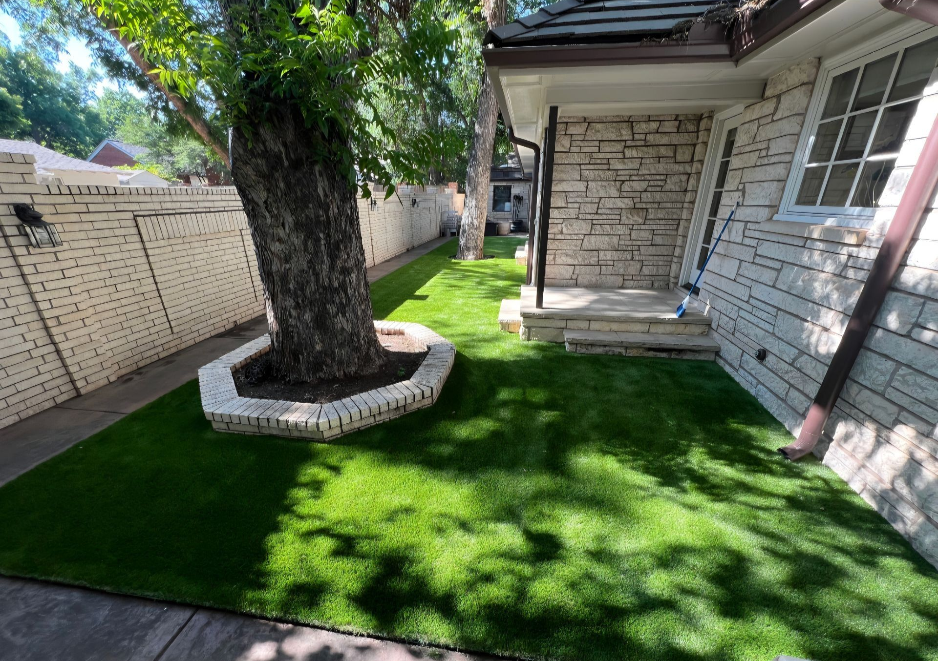 Narrow backyard with grass, gravel borders, tall trees, a fence, and a small garden bed in bright sunlight