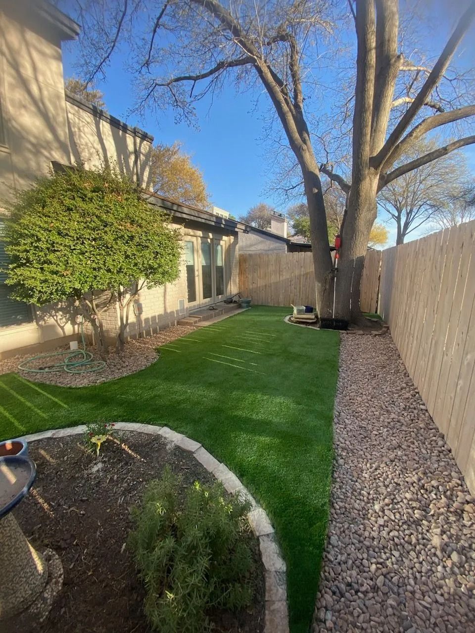 Narrow backyard with artificial grass, bare trees, gravel border, fence, and a small garden bed in bright sunlight