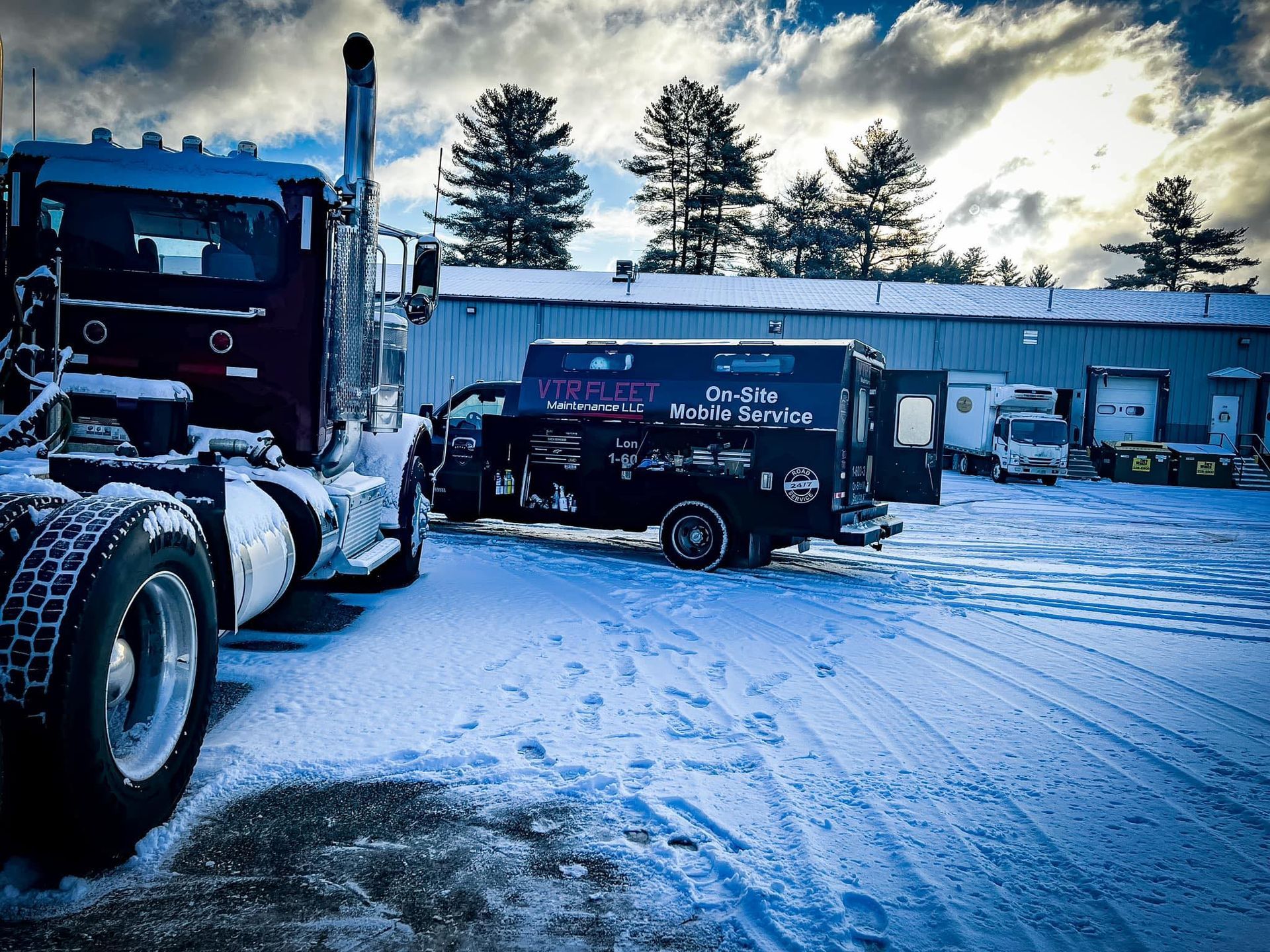 Semi-truck and service van parked in a snow-covered lot. Building and trees in the background under cloudy skies.
