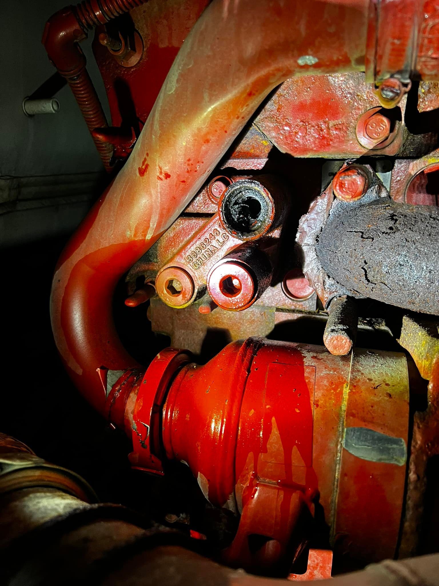 Close-up of a corroded red engine component with several open holes and fluid drips.