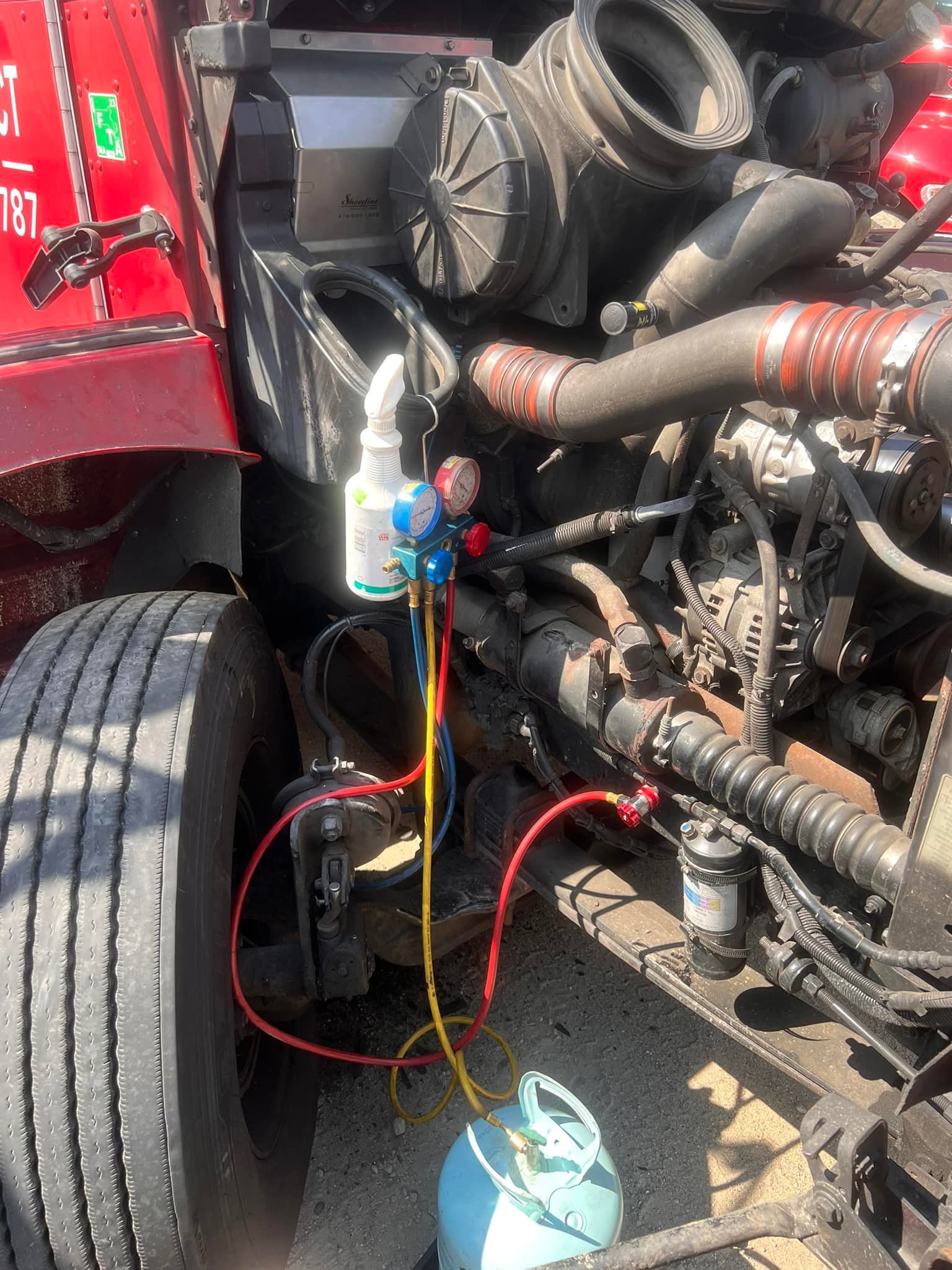 A semi-truck engine bay with gauges and refrigerant tank connected, likely for A/C repair.
