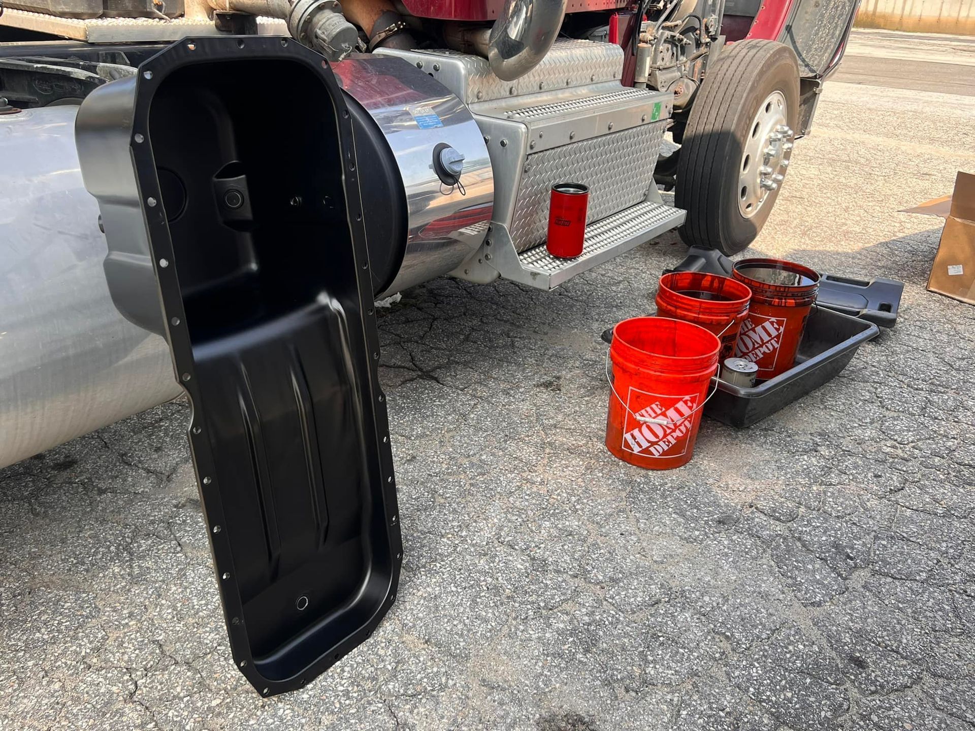 A large black oil pan and red Home Depot buckets are on gravel next to a truck.