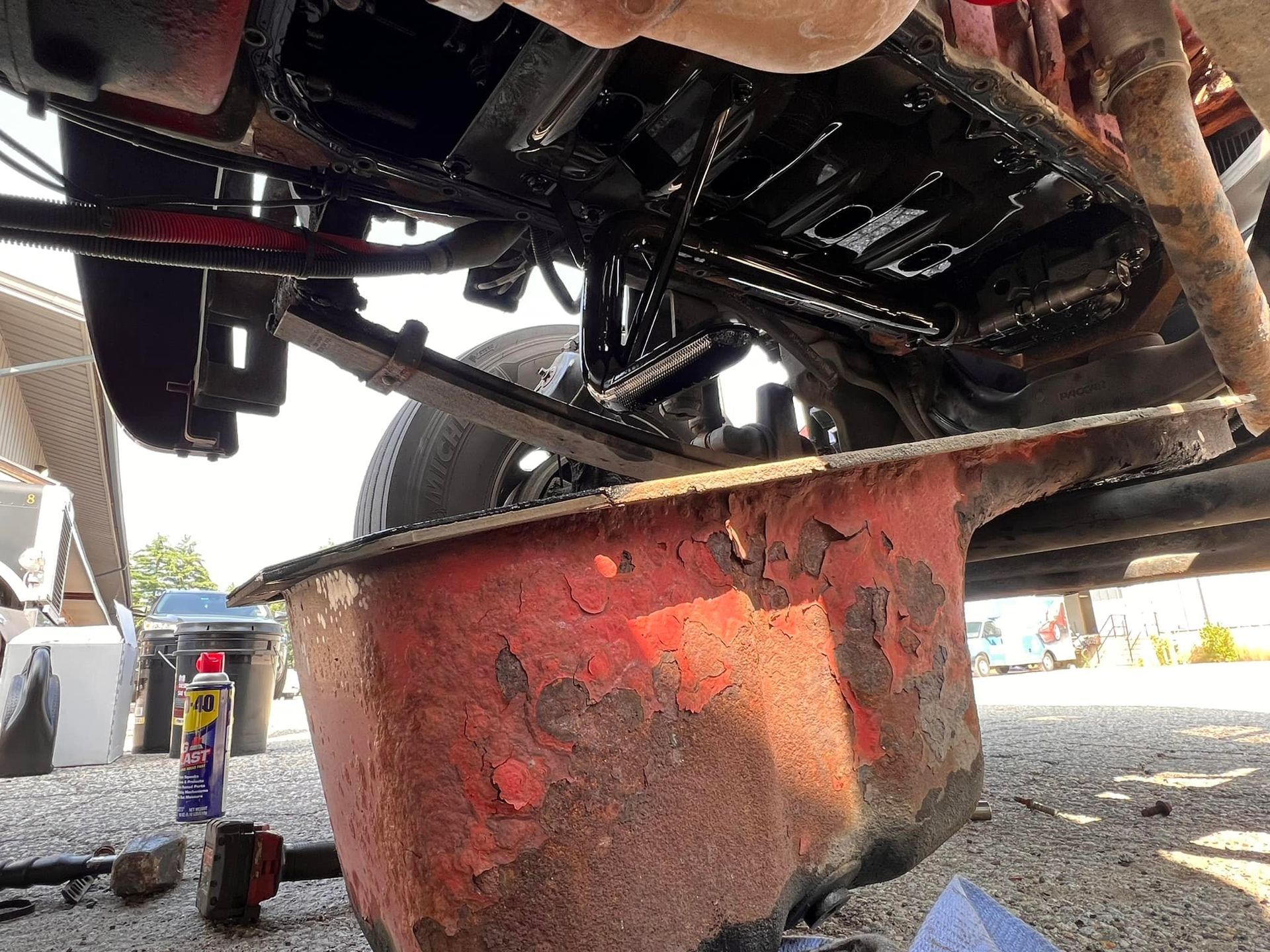 Underneath a vehicle, a rusty oil pan collects dark oil during a car maintenance activity.