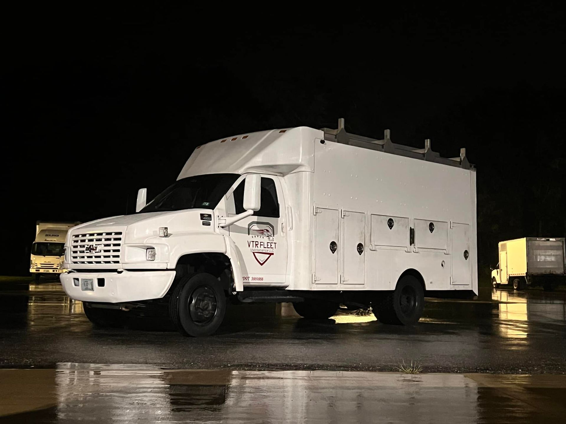 White work truck parked on a wet asphalt lot at night. Black wheels, side storage compartments, roof racks.