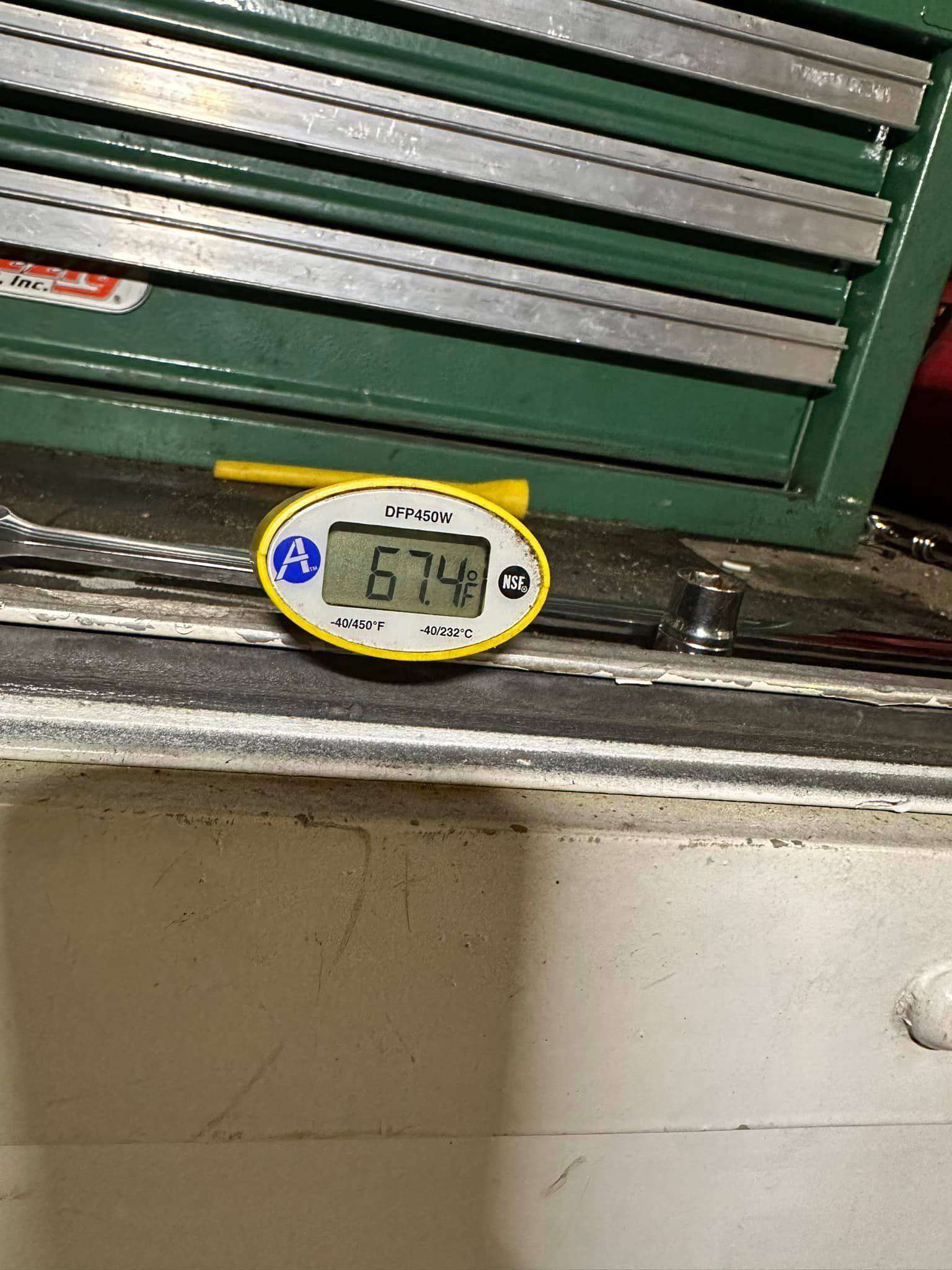 Digital thermometer reads 67.4 degrees; yellow casing on a dusty surface with a green cabinet in the background.