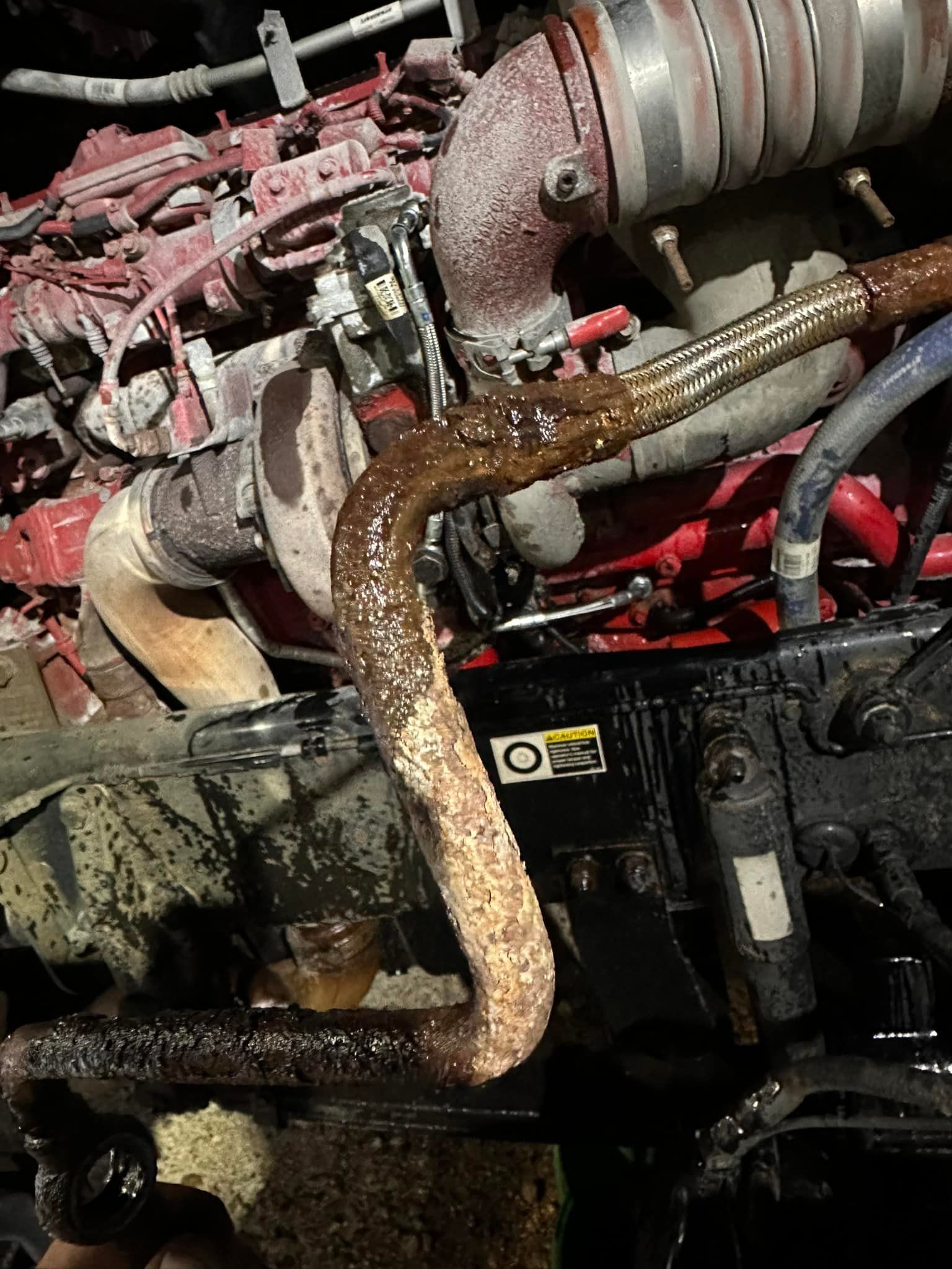 Rust-covered pipe attached to a truck engine, covered in oil and debris, with red and black components.