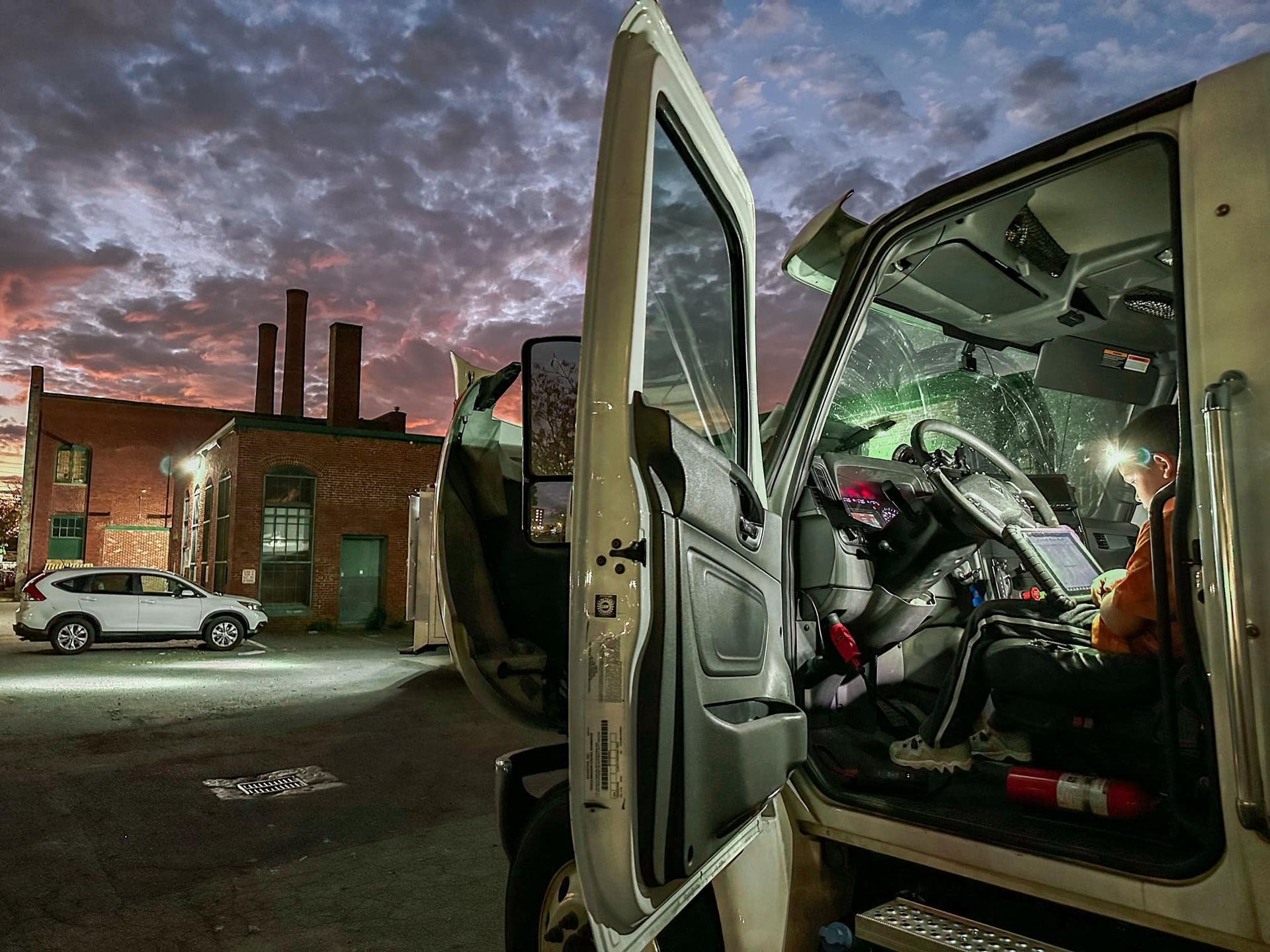 Truck cabin, door open, driver inside at dusk. Red brick buildings and car in the background, blue and orange sky.