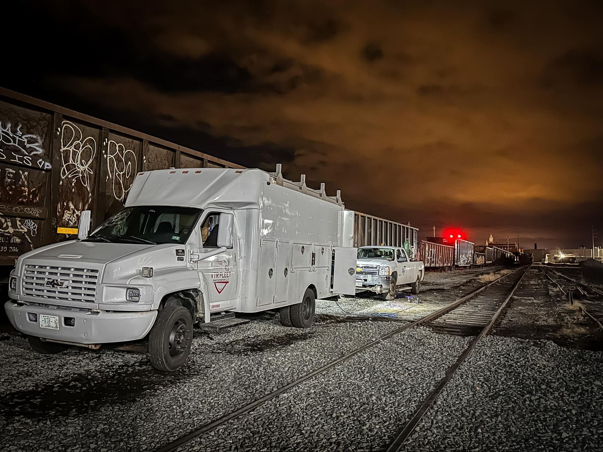 A large white utility truck and trailer on train tracks at night, with a train car in the background.