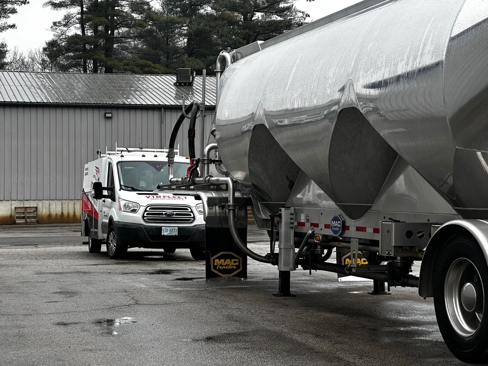 White van and silver trailer connected at a loading dock, black hoses attached.