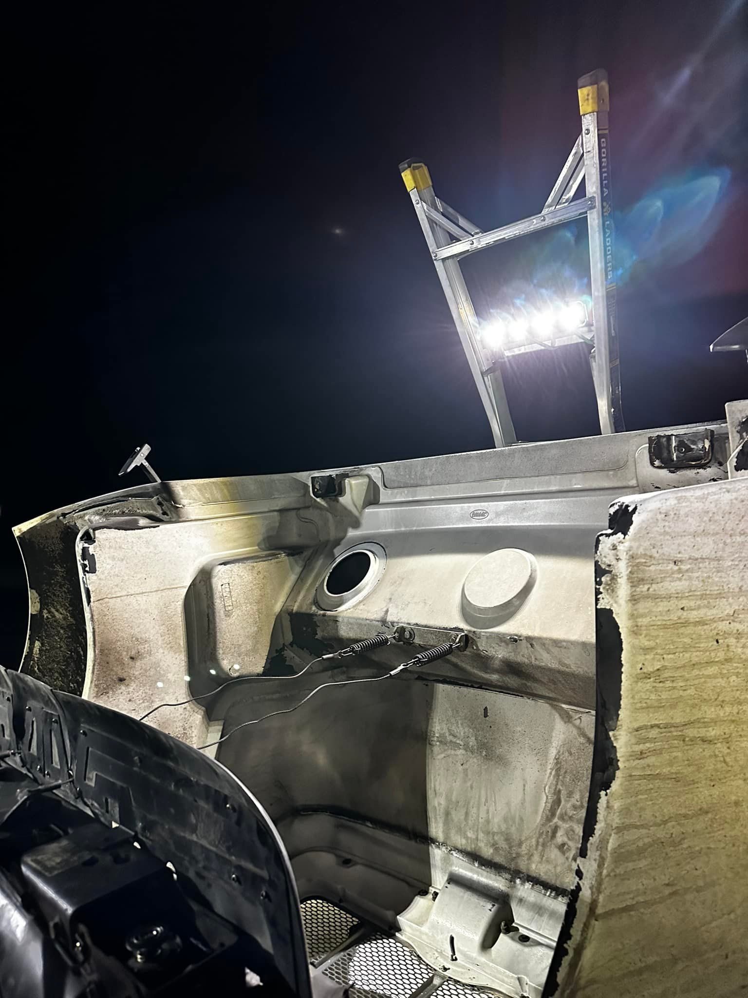 A damaged car frame illuminated at night with a metal ladder and bright lights.
