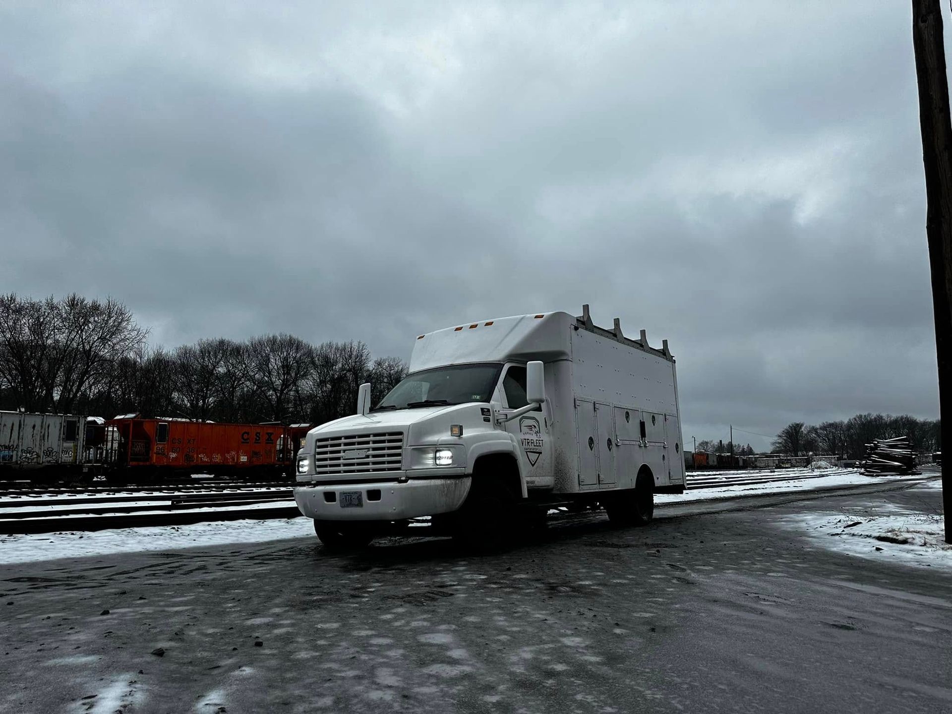 White utility truck on snow-covered asphalt, railway tracks and cloudy sky in background.