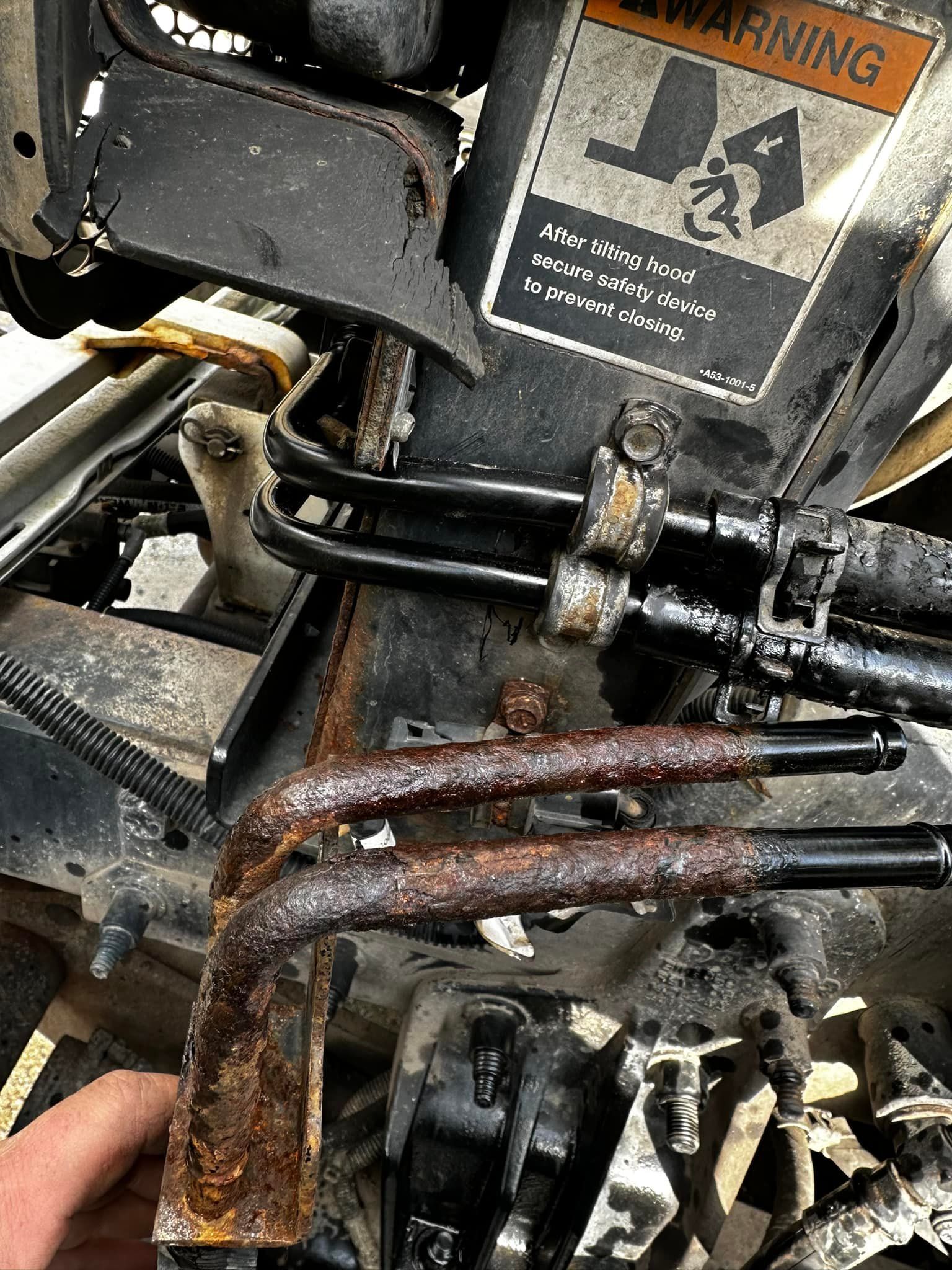 Rusty metal tubes on a vehicle frame, possibly part of a hydraulic system. Warning label visible.