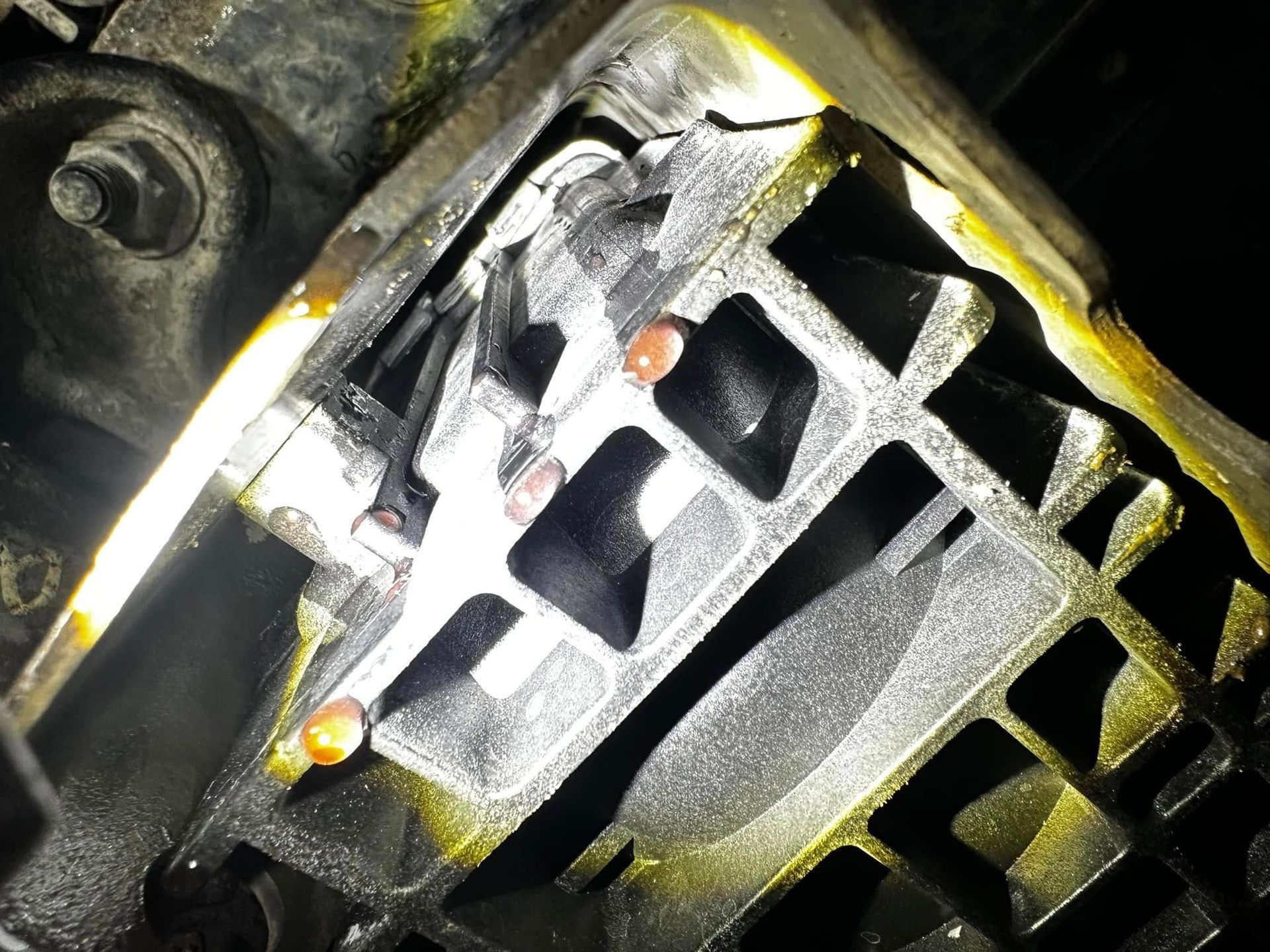 Close-up of a partially disassembled, oil-stained vehicle engine component. Yellow oil residue covers the metal structure.