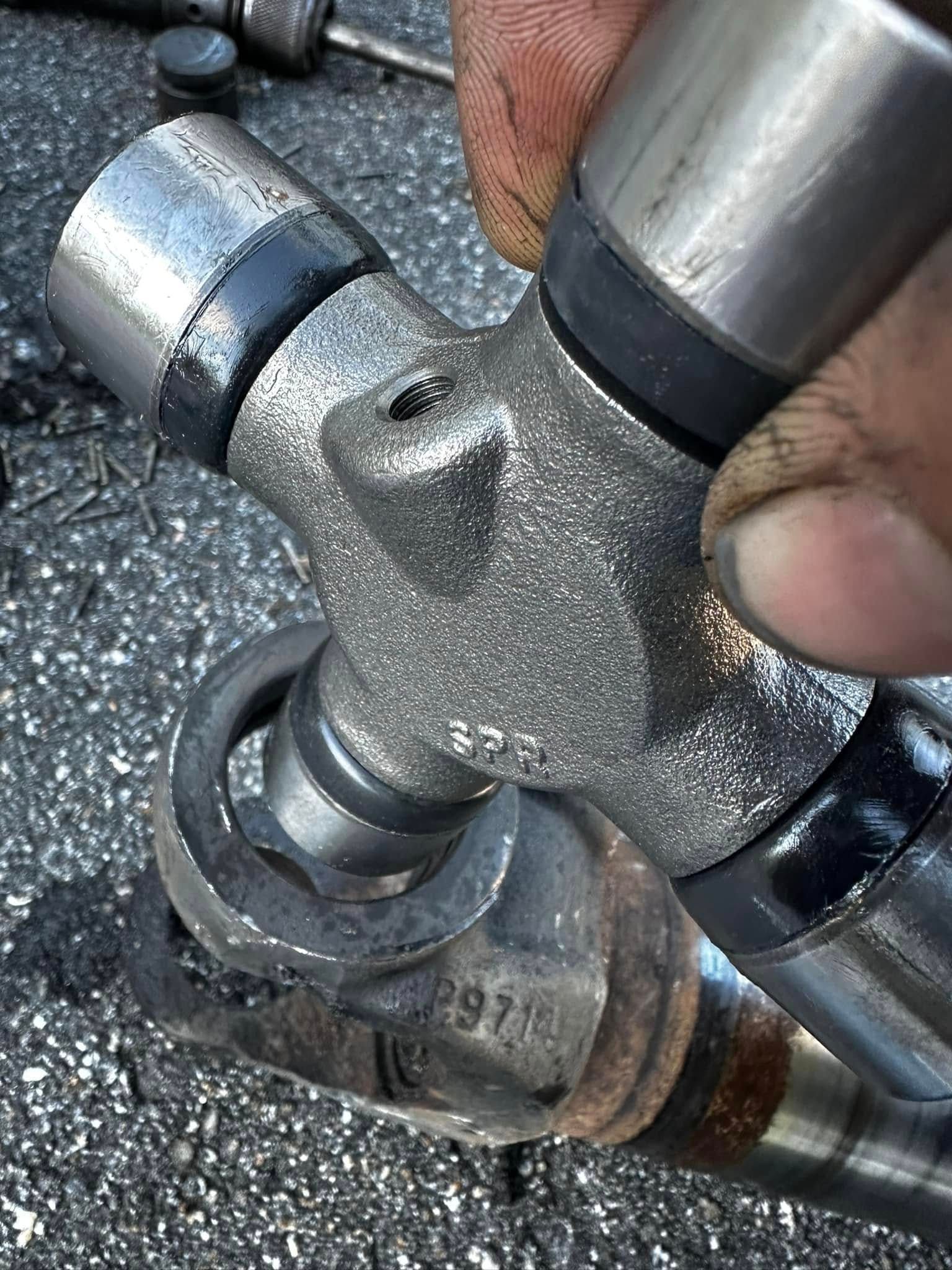 Close-up of a U-joint assembly, held by a hand. The joint is silver and black, with a rusty component below.