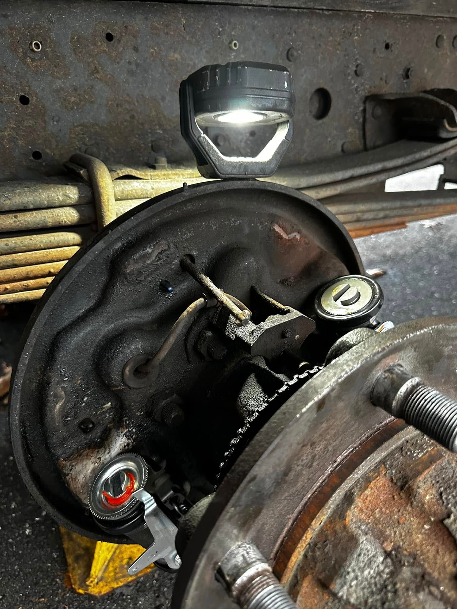Close-up of a truck's rear brake assembly with a work light attached, tools, and a rusty truck frame.