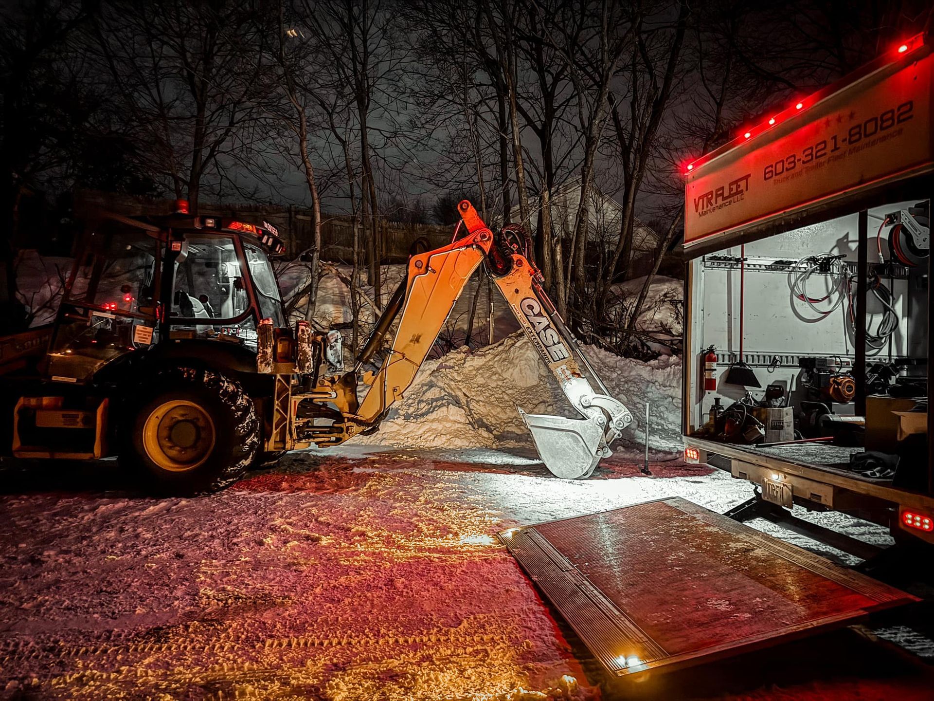 Backhoe loading snow into a trailer at night. Yellow machinery, white snow, trees in background.